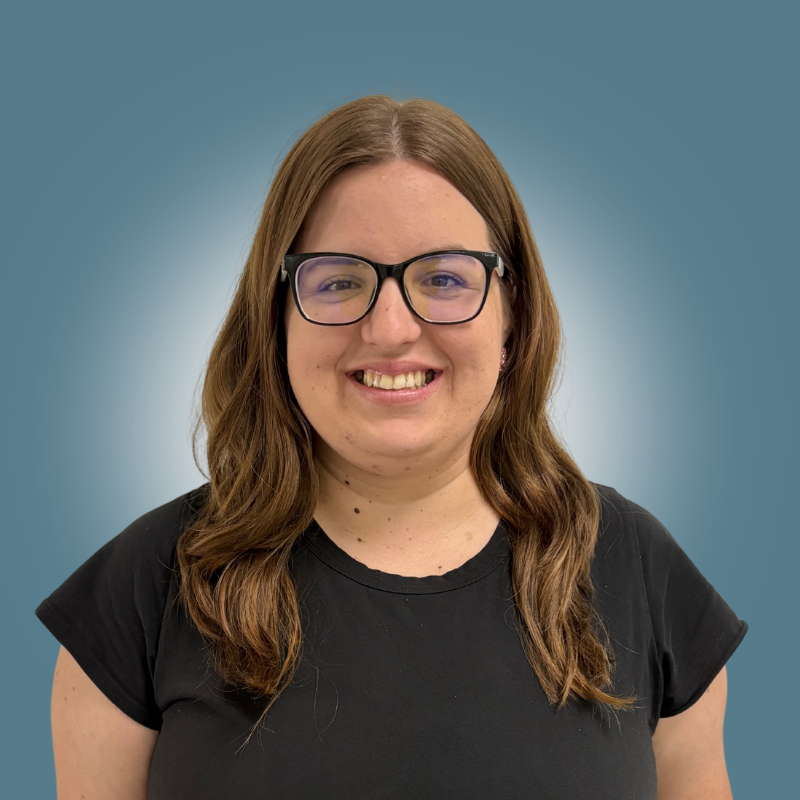 A young woman with brown hair, glasses, and a black t-shirt smiling at the camera, photo taken against a blue background.