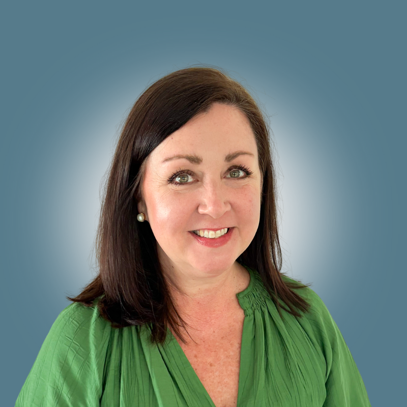 A woman with shoulder-length dark brown hair, fair skin, and blue eyes smiling, wearing a green blouse and pearl earrings, against a blue background.