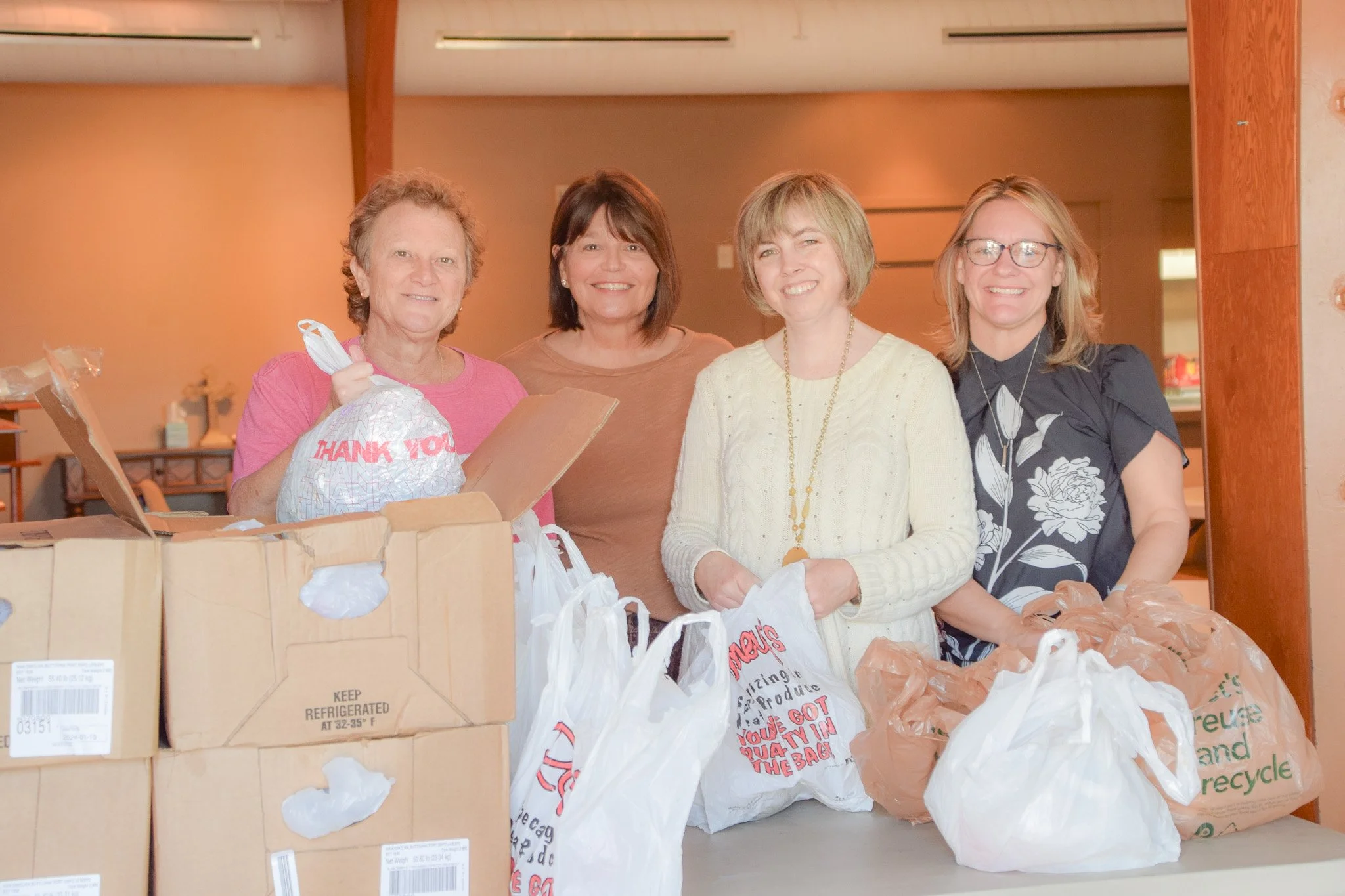 Four women standing behind a table with bags and boxes of groceries, smiling at the camera.