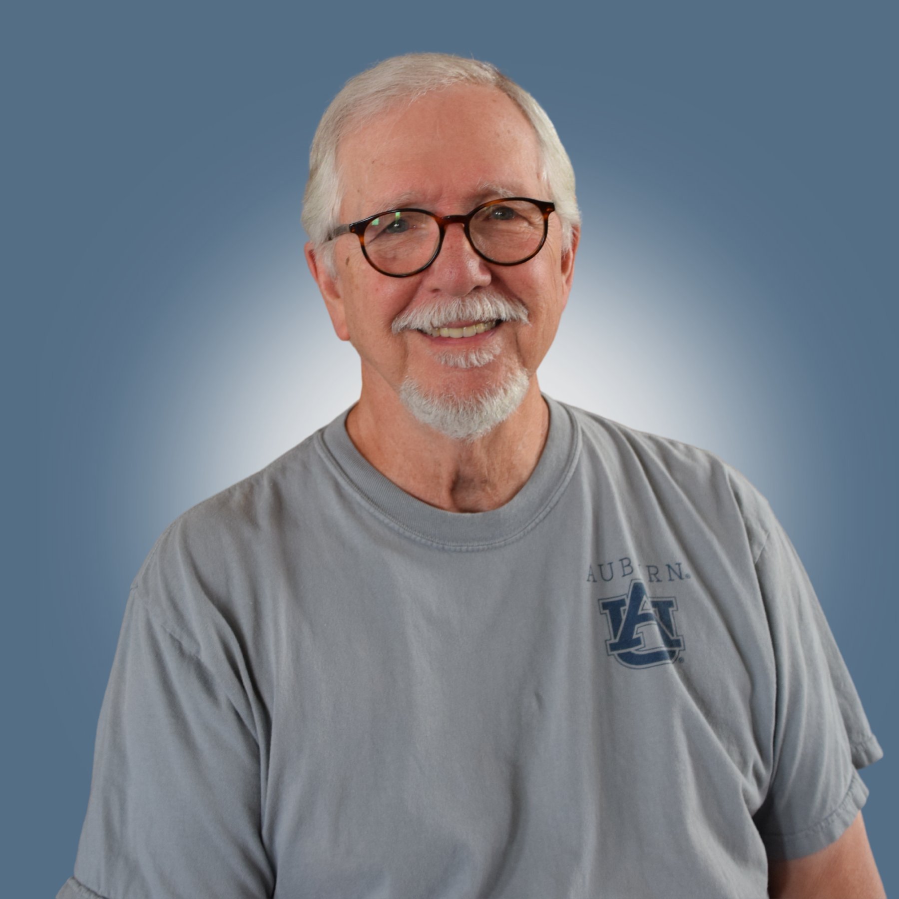 An elderly man with glasses and a beard smiles at the camera, wearing a gray t-shirt with Auburn University logo, against a blue background.
