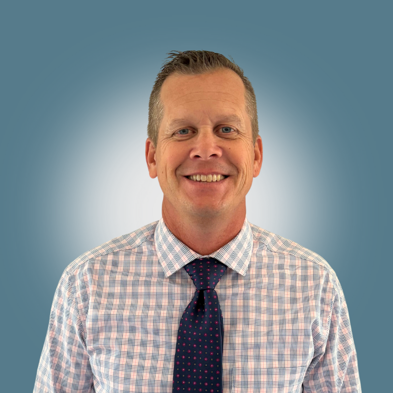 A smiling man wearing a checkered shirt and a dark, dotted tie, standing against a gradient blue background.