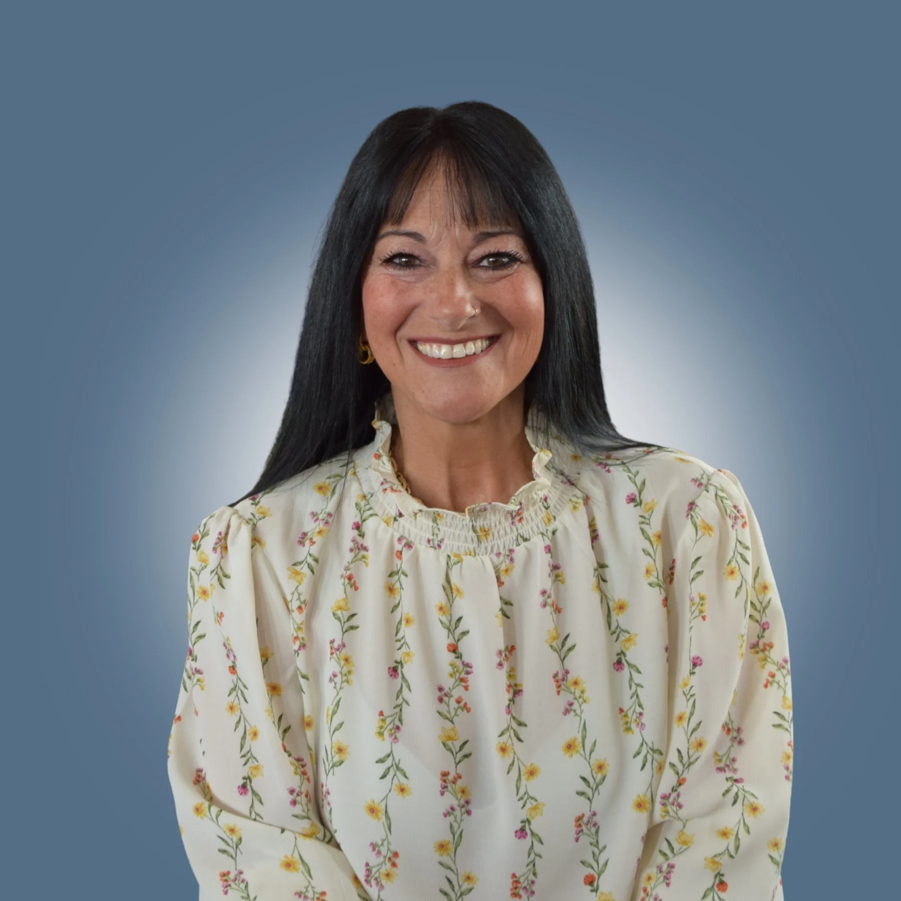 A woman with long black hair, smiling, wearing a cream-colored blouse with a floral pattern and a high ruffled neckline, against a blue background.