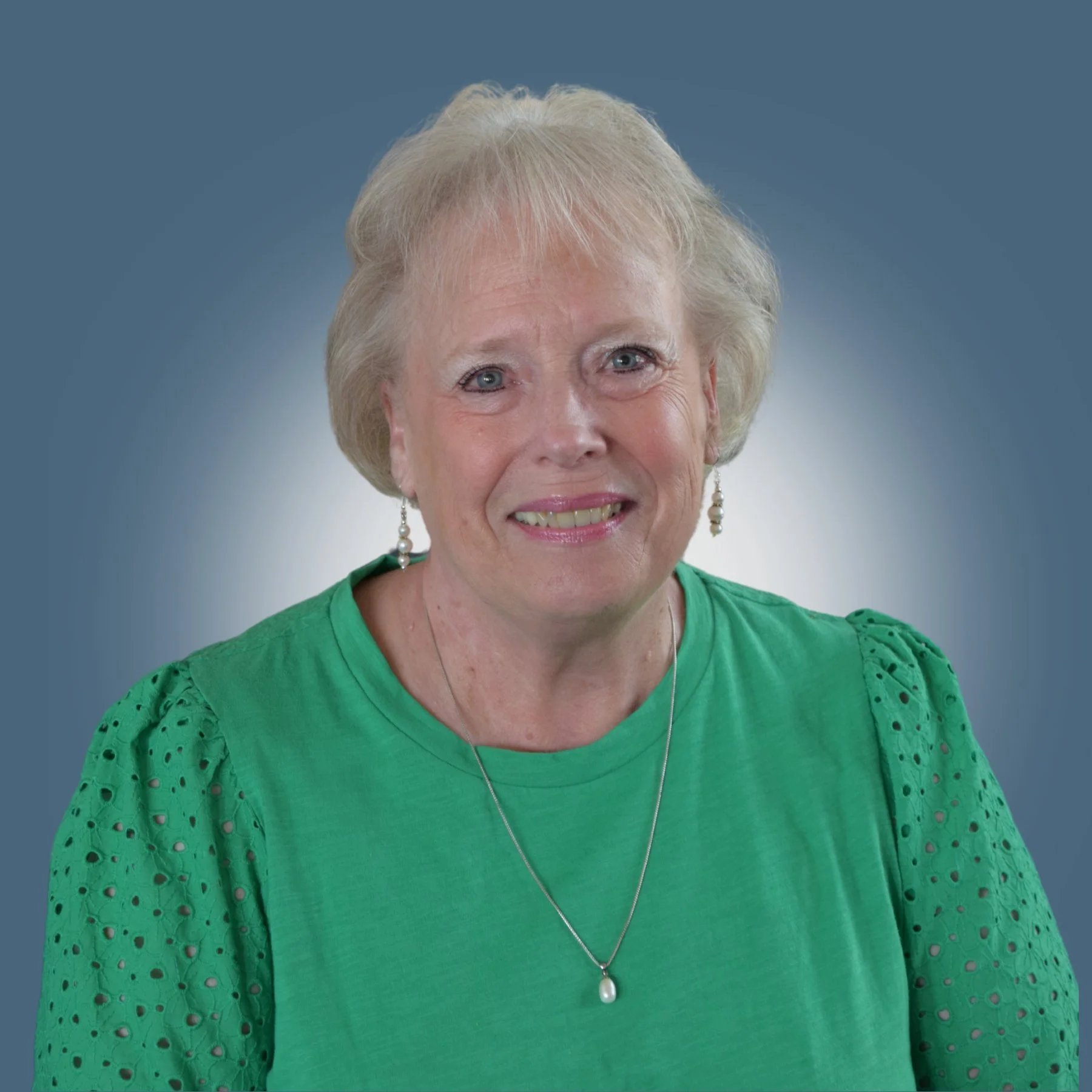 A portrait of an elderly woman with short blonde hair, wearing a green top with eyelet sleeves, pearl earrings, and a pearl necklace, smiling against a blurred blue background.