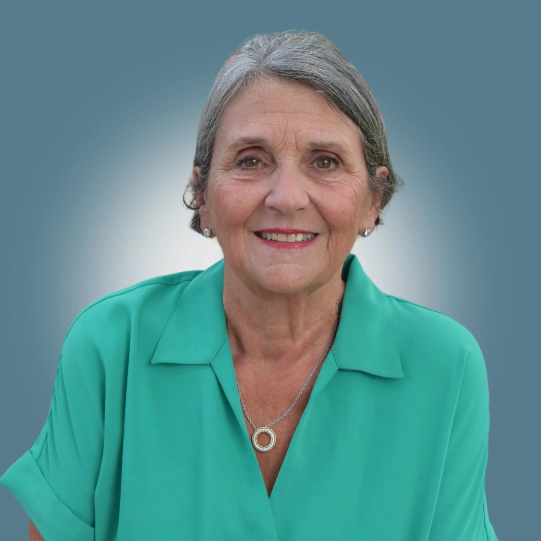 Portrait of an older woman with short gray hair, wearing a teal blouse and jewelry, smiling at the camera.