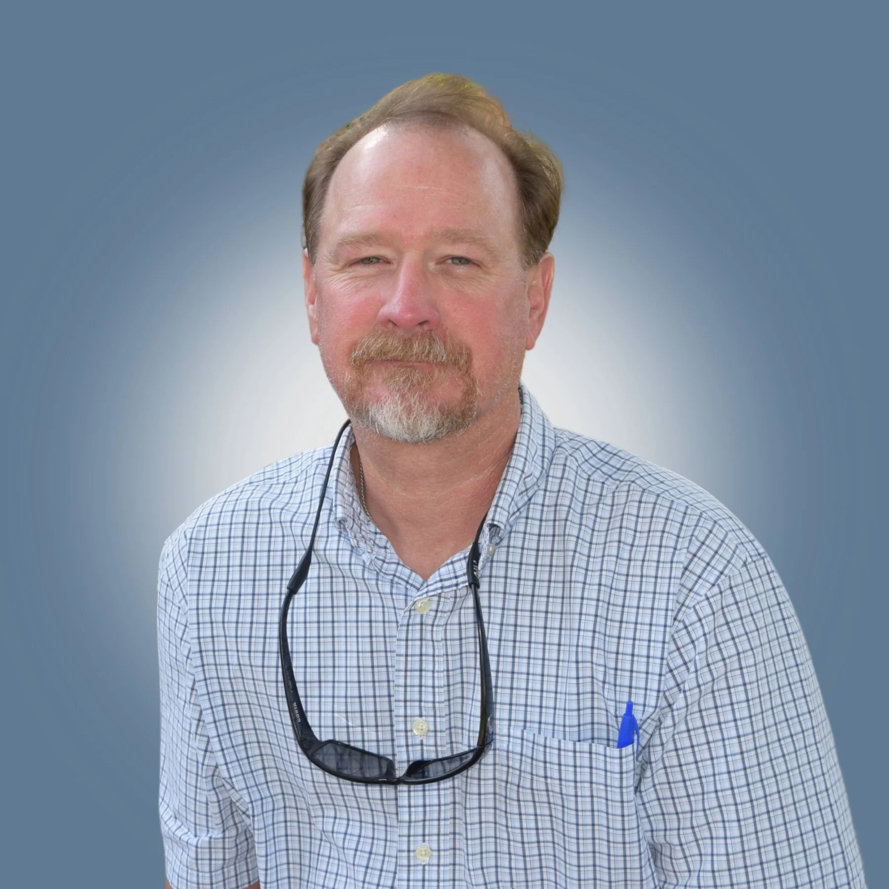 A middle-aged man with a beard and glasses hanging around his neck, wearing a checkered shirt and a blue pen in his pocket, posing against a plain background.