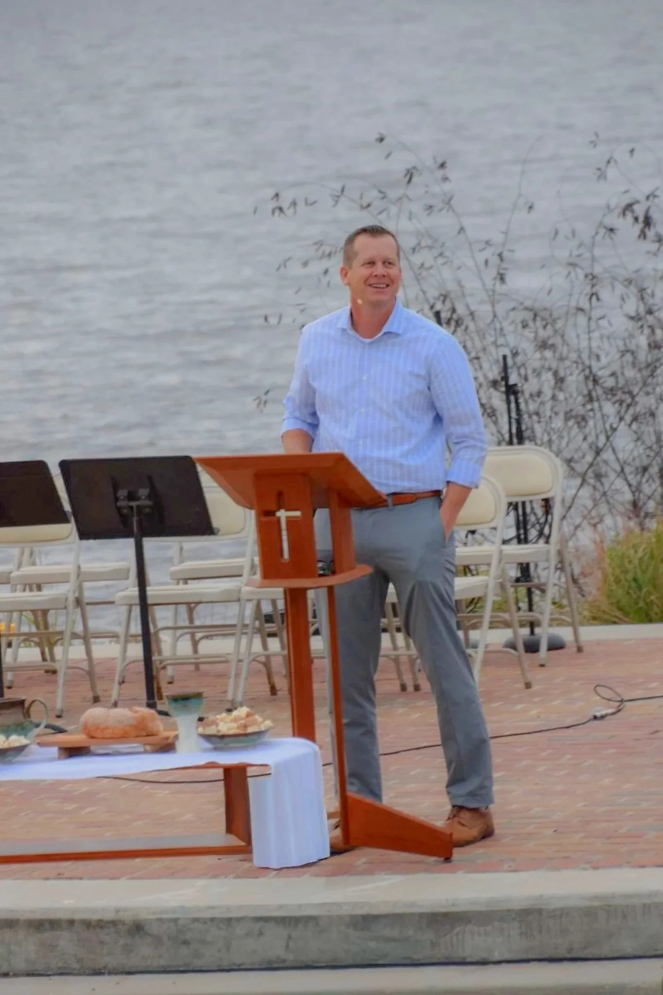 A man stands outdoors near a lake, behind a wooden podium with a cross on it, wearing a striped shirt and gray pants, smiling.
