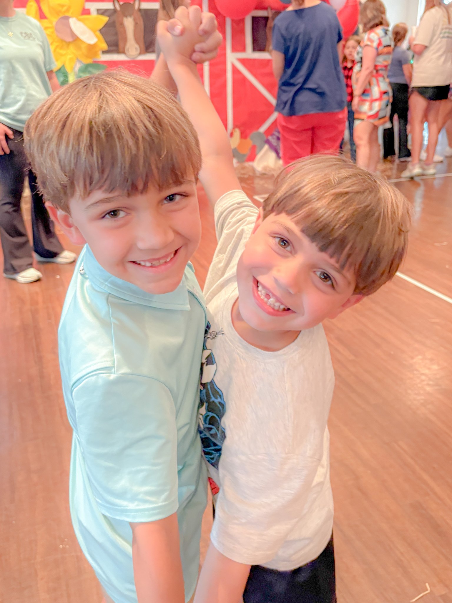 Two young boys with brown hair smiling and holding hands at a lively event, surrounded by other children and adults in a decorated indoor space.