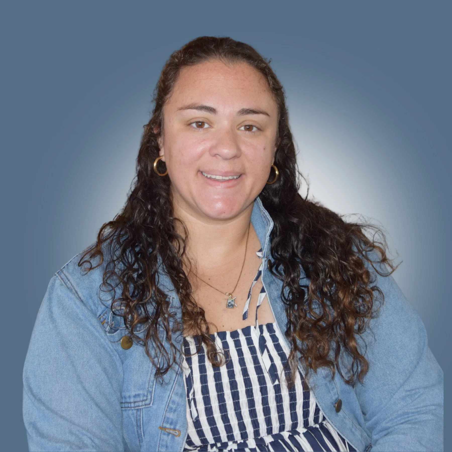 A woman with curly brown hair wearing a denim jacket and striped dress, smiling against a blue gradient background.