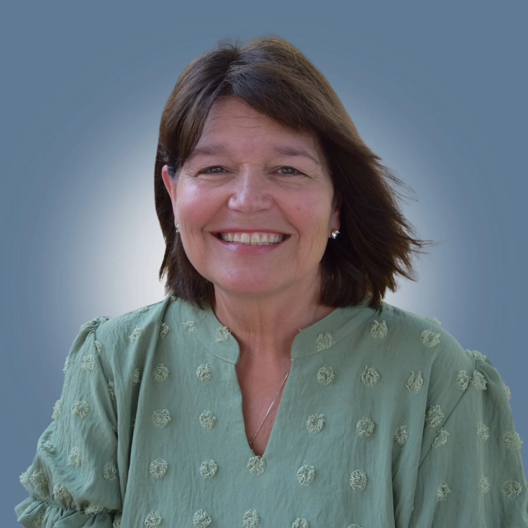 A smiling middle-aged woman with shoulder-length brown hair, wearing a light green blouse with embroidered circular patterns, standing against a soft blue background.