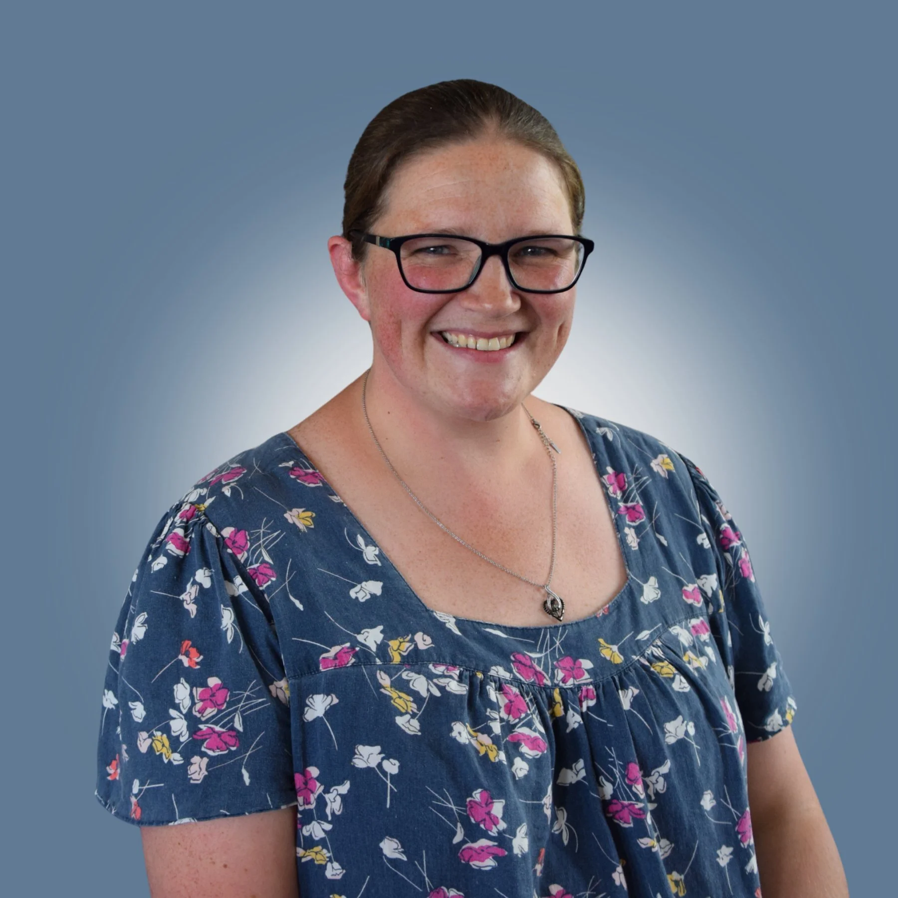 Portrait of a woman with glasses, smiling, wearing a floral dress and a necklace against a blue gradient background.