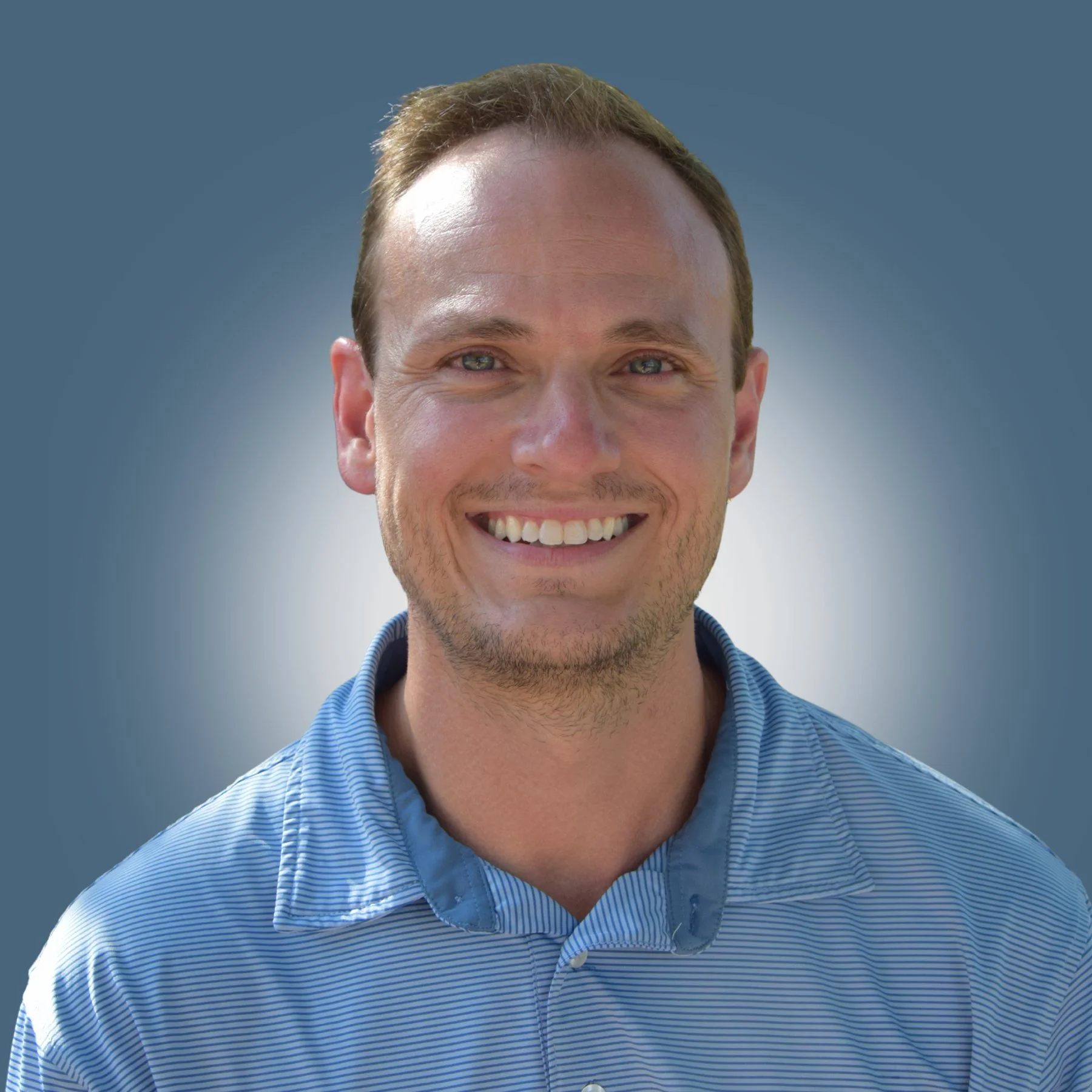 A smiling man with short brown hair and a light beard, wearing a blue striped collared shirt, standing against a blue-gray gradient background.