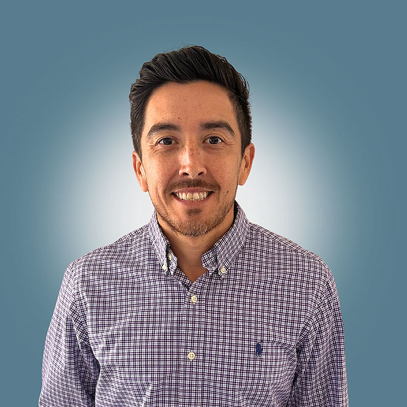A smiling man with short dark hair and a beard, wearing a checkered button-up shirt, standing against a blue gradient background.