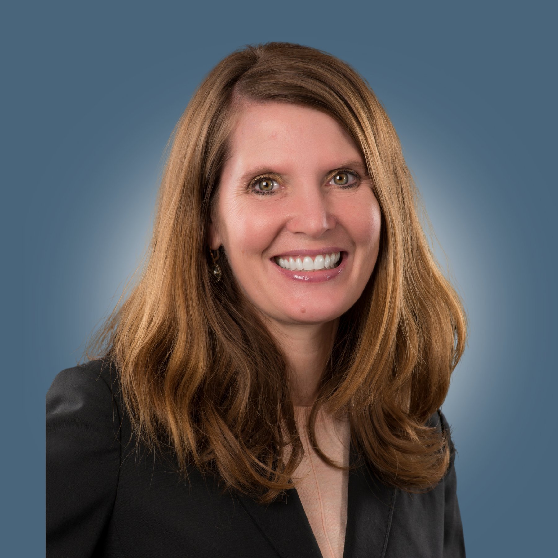 Headshot of a smiling woman with long, wavy brown hair, wearing a black blazer, against a blue background.