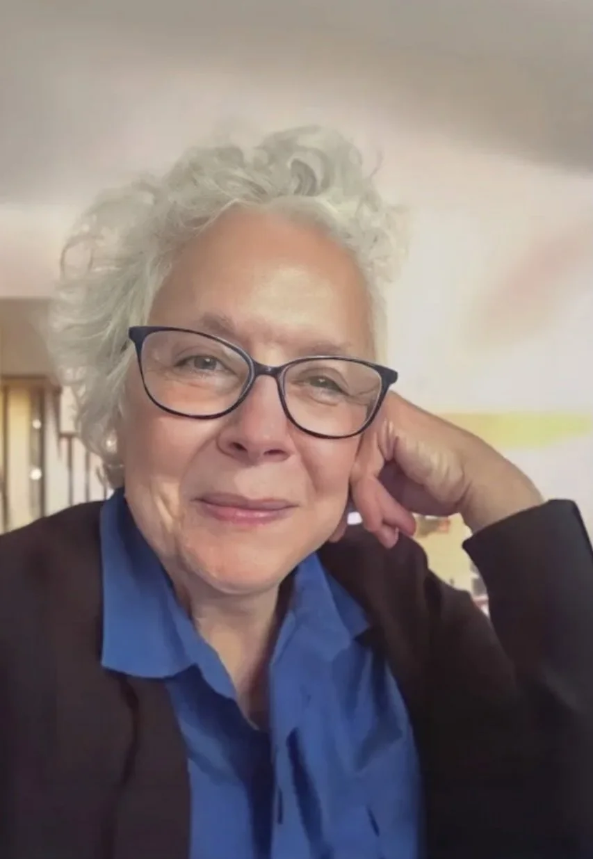 A woman with curly gray hair and glasses, wearing a blue shirt and a dark cardigan, is sitting indoors resting her head on her hand and smiling gently.