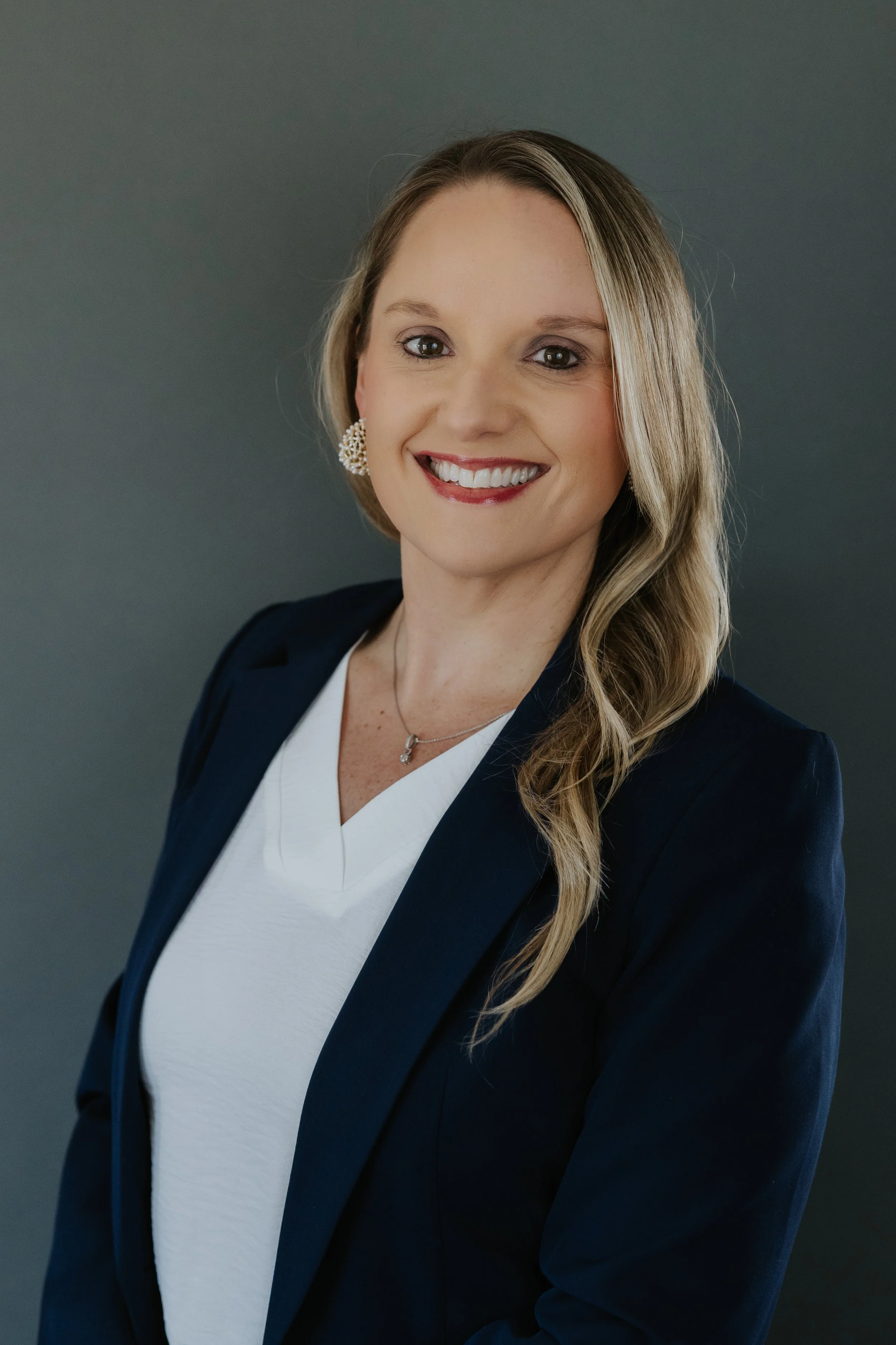 Professional woman with blonde hair, wearing a navy blazer, white blouse, and jewelry, smiling against a gray background.