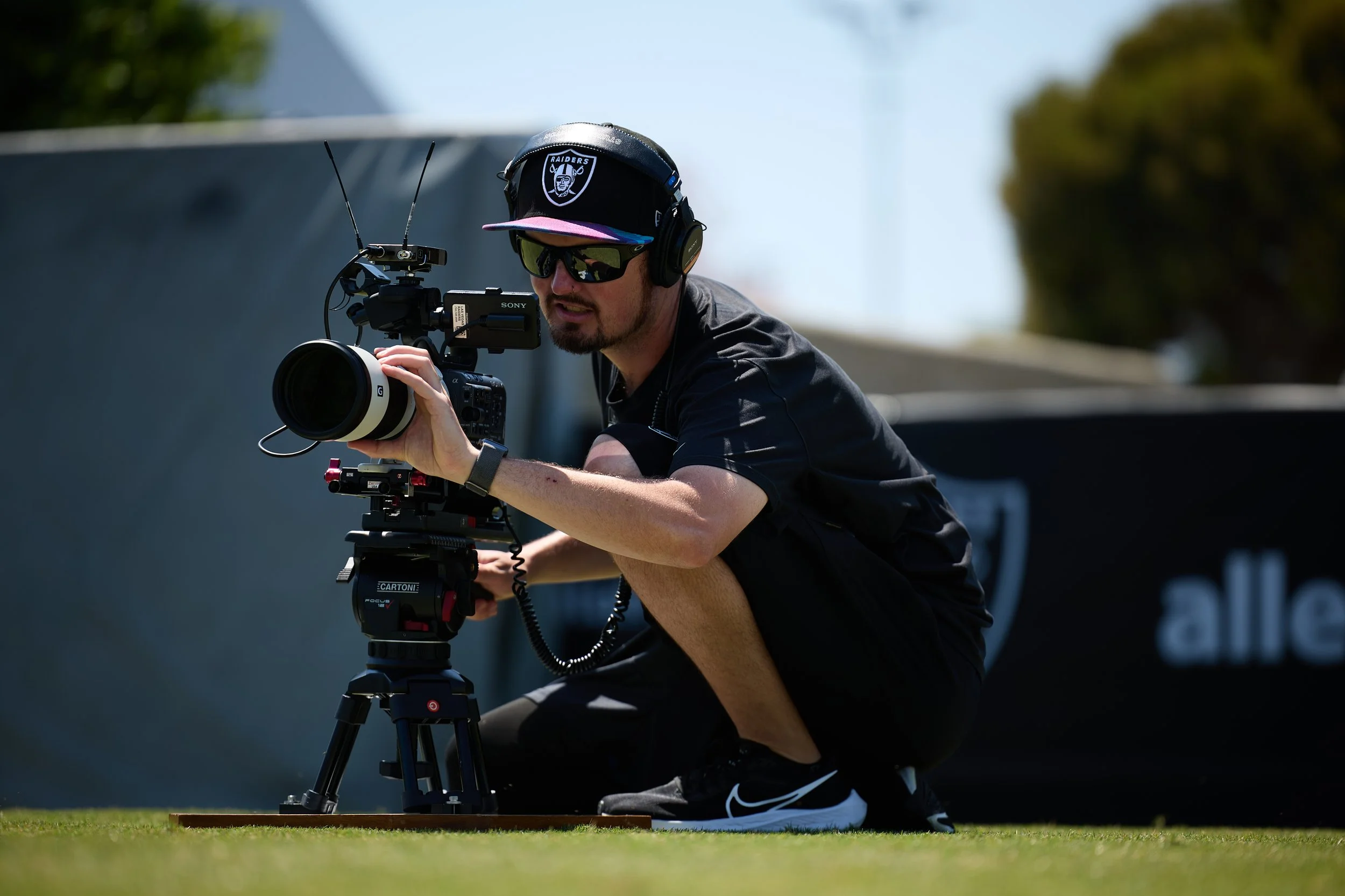 Sam Freed operating a professional video camera outdoors, wearing sunglasses, a black Las Vegas Raiders cap, and headphones, crouching on a sunny day shooting Las Vegas Raiders football practice.