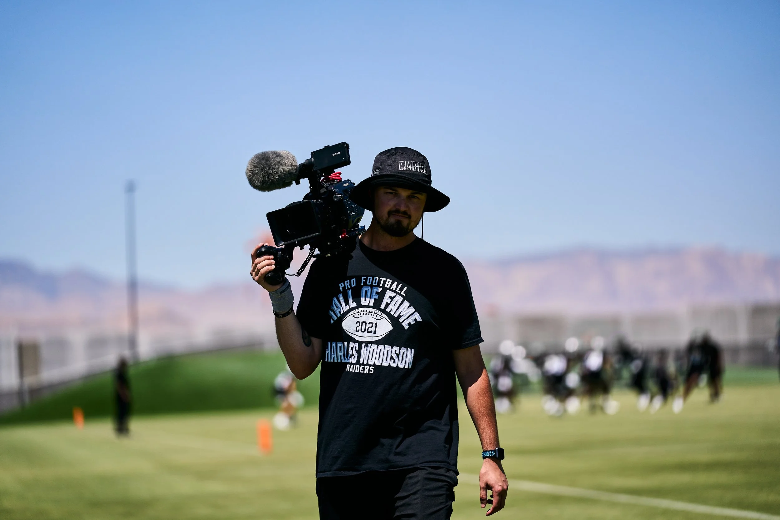 A man is walking outdoors on a sunny day, carrying a professional video camera on his shoulder. He is wearing a black hat, a black t-shirt with text, and a black wristwatch.