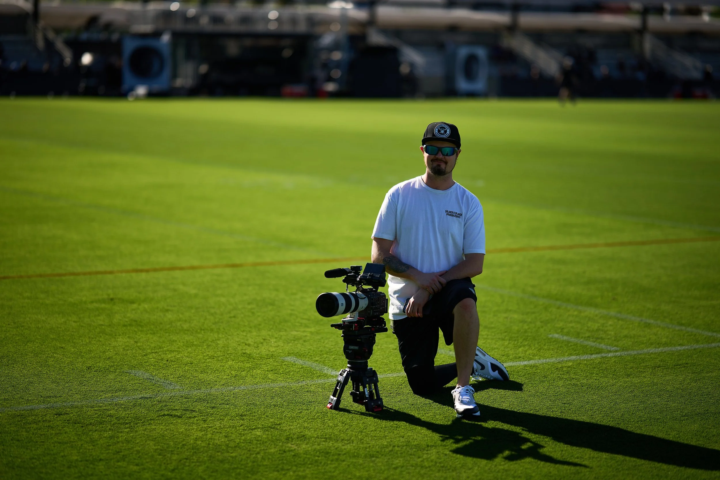 A man kneeling on one knee on a grassy sports field, holding a professional camera setup on a tripod, wearing sunglasses, a cap, a white t-shirt, and shorts.