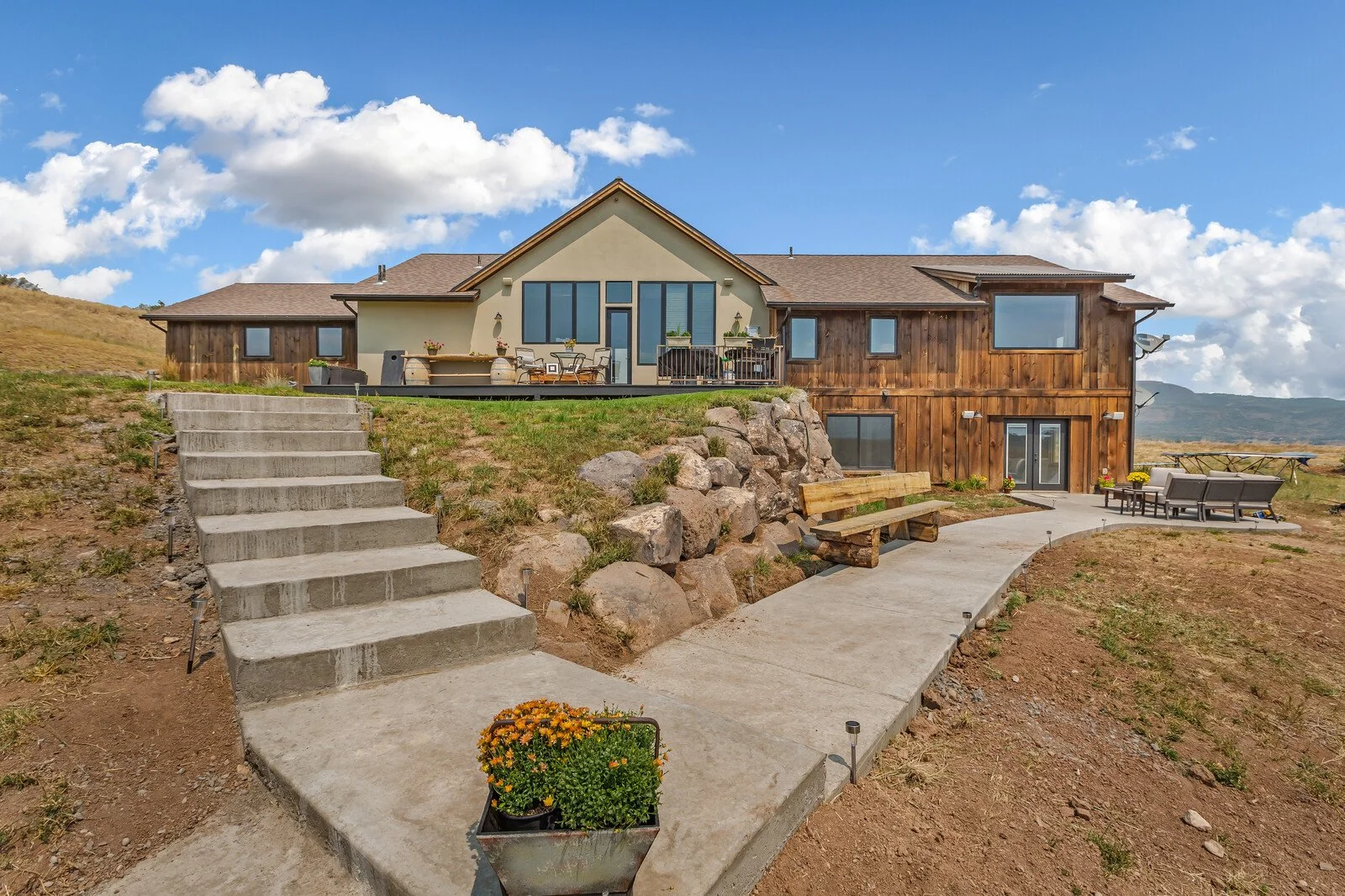 A modern house with a mix of beige and wood exterior, large windows, and a spacious outdoor patio with seating, surrounded by a concrete pathway, stairs, and rocky landscaping under a blue sky with clouds.