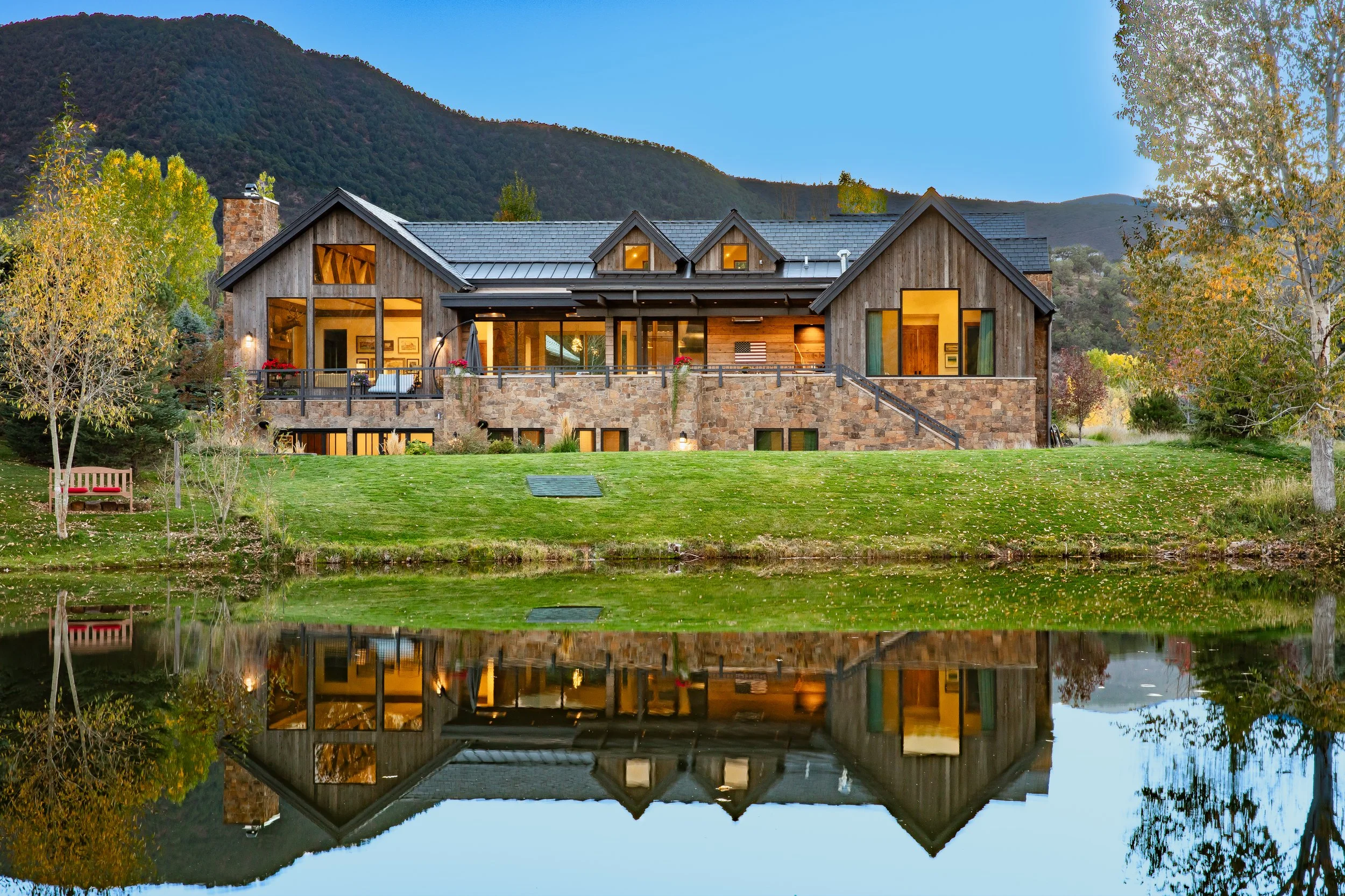 A large house with a wooden and stone exterior, situated on a grassy hillside with trees, reflected in a calm body of water in the foreground, mountains in the background, and a clear blue sky.