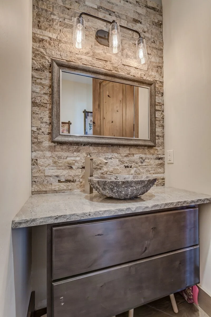 Bathroom vanity area with a stone vessel sink, a rectangular mirror, stone backsplash, and a modern light fixture with three glass bulbs.