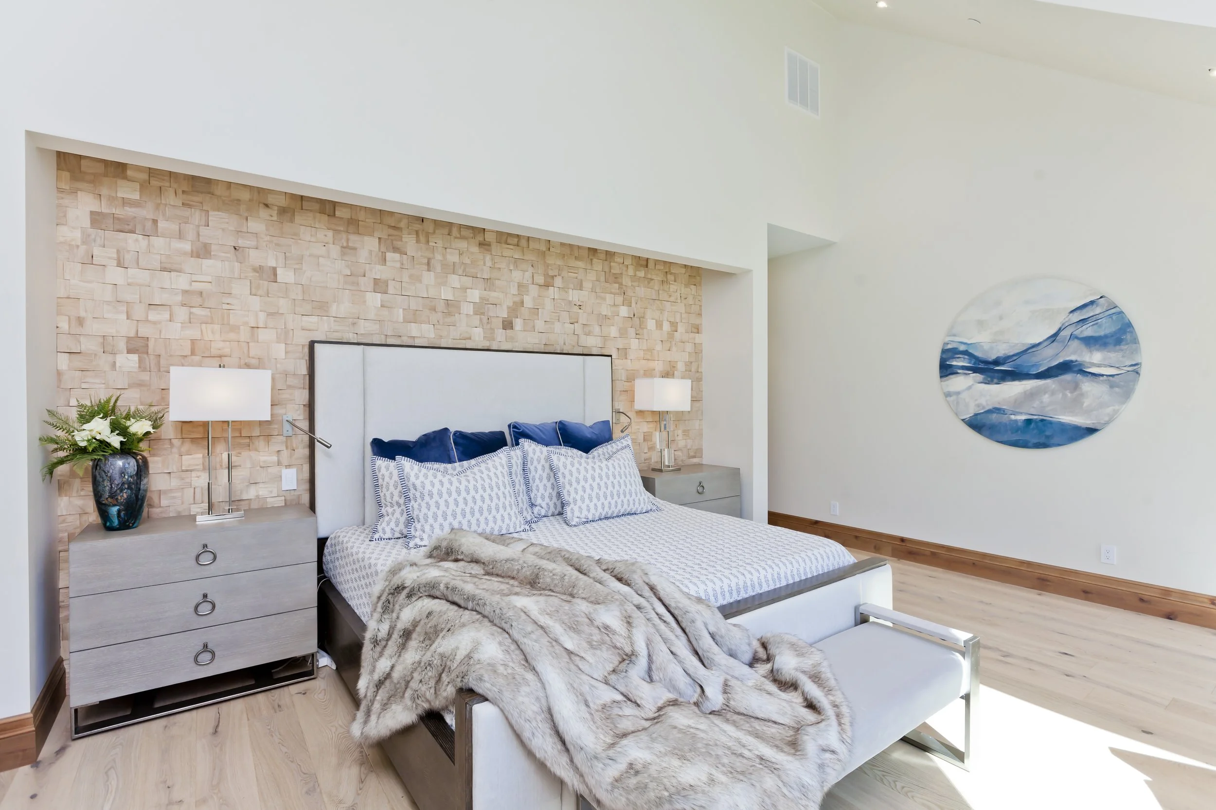 A modern bedroom with a white bed, gray nightstands with lamps, blue pillows, a fur throw, a pinkish brick accent wall behind the bed, a circular abstract blue and white wall art, and light wood floors.