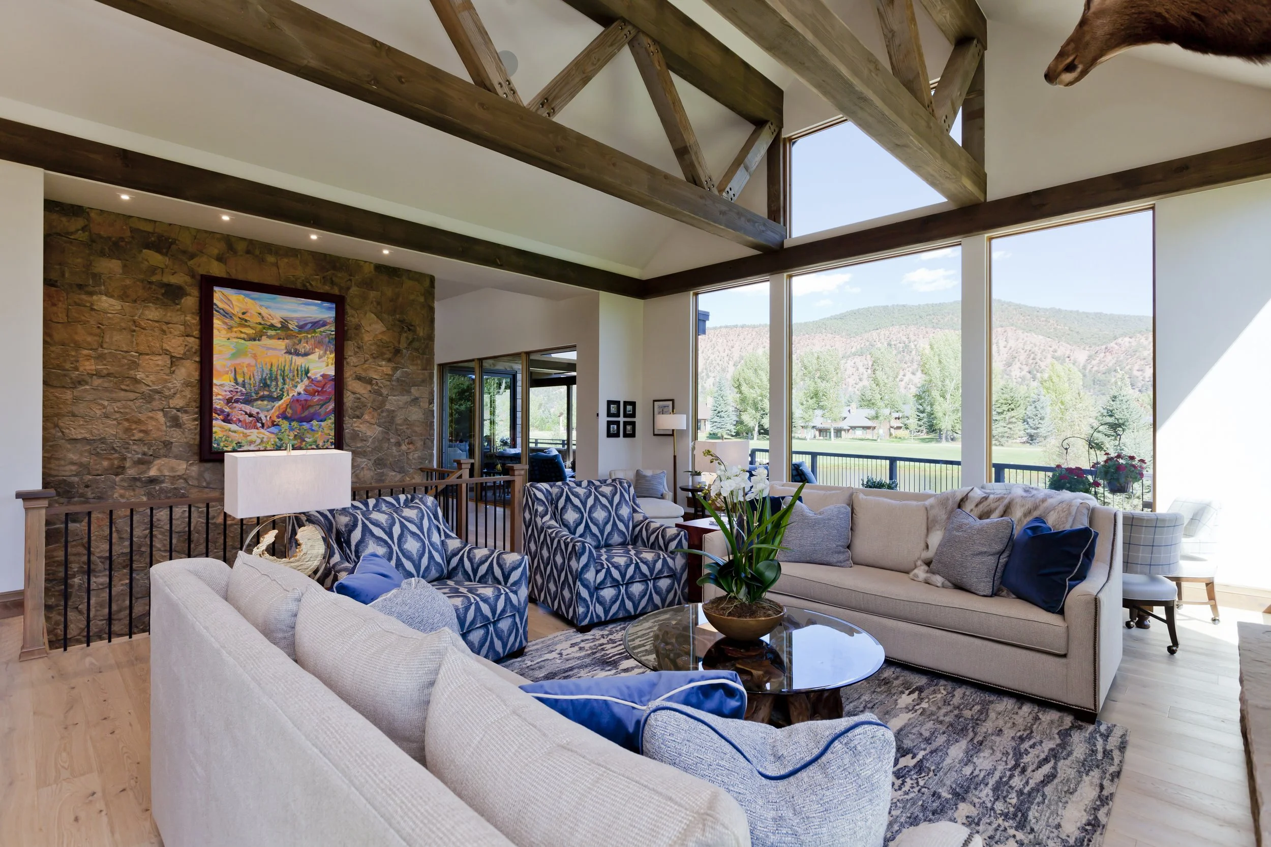 Living room with large windows showing mountains and trees, beige and blue furniture, a coffee table with a plant, wall art, and decorative pillows.