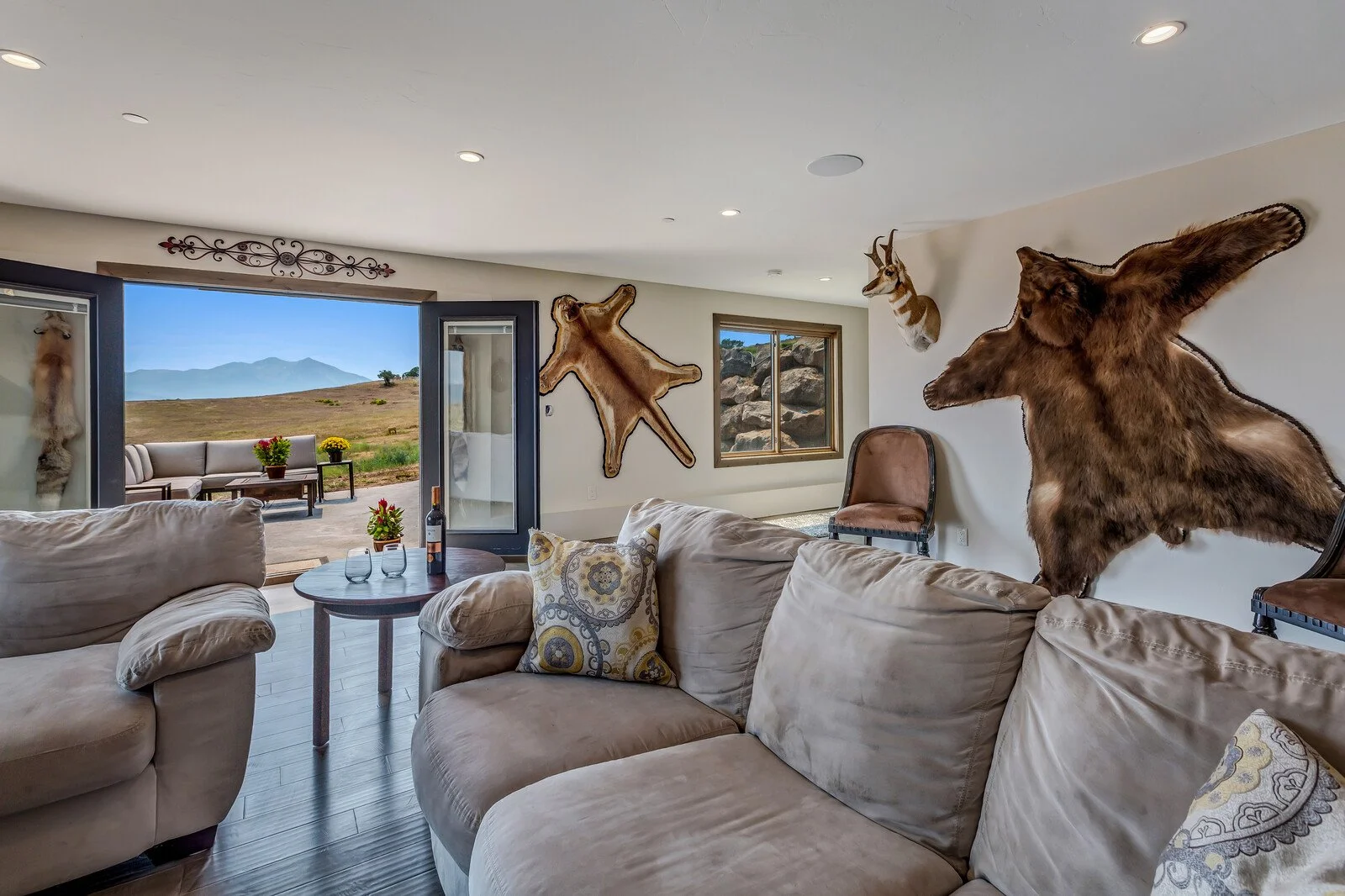 Living room with animal hide wall decorations and open sliding doors to outdoor patio with mountain view, beige sofa, pillows, small round table, and chairs.