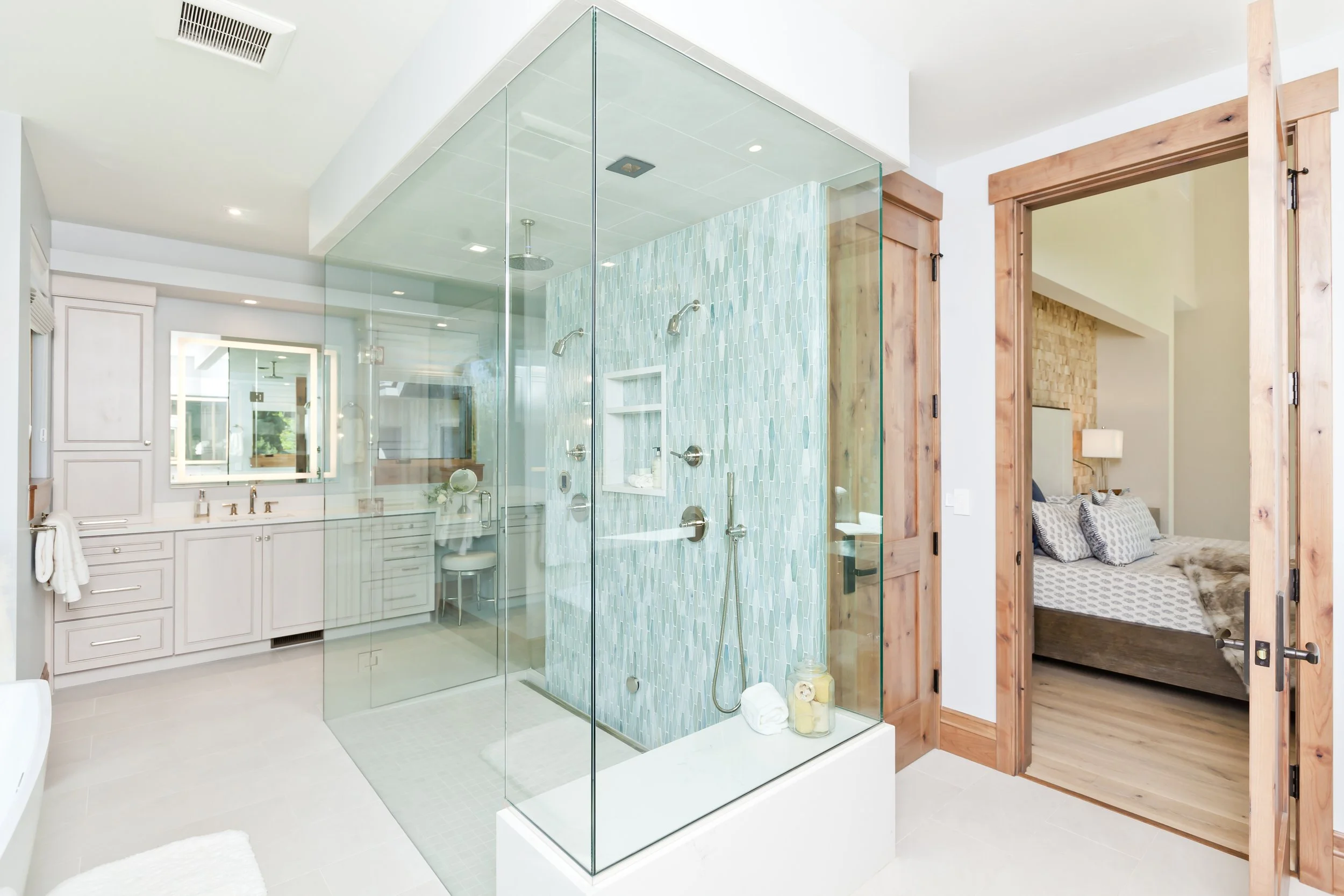 Modern bathroom with a glass shower enclosure, light blue mosaic tiles, white vanity with mirror, and a doorway to a bedroom with wooden trim.
