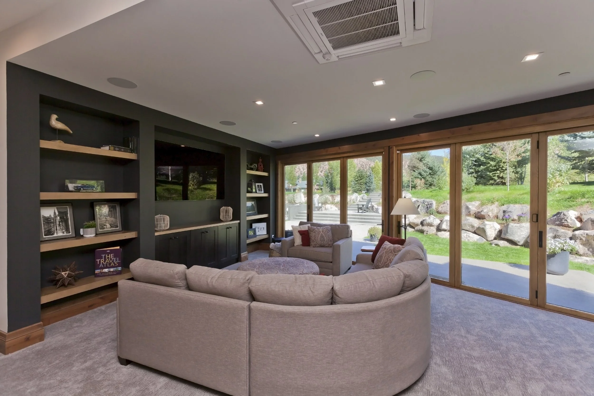 Living room with large sliding glass doors, beige sectional sofa, and built-in wall shelves with decor and pictures. Outdoors visible include rocks, trees, and greenery.