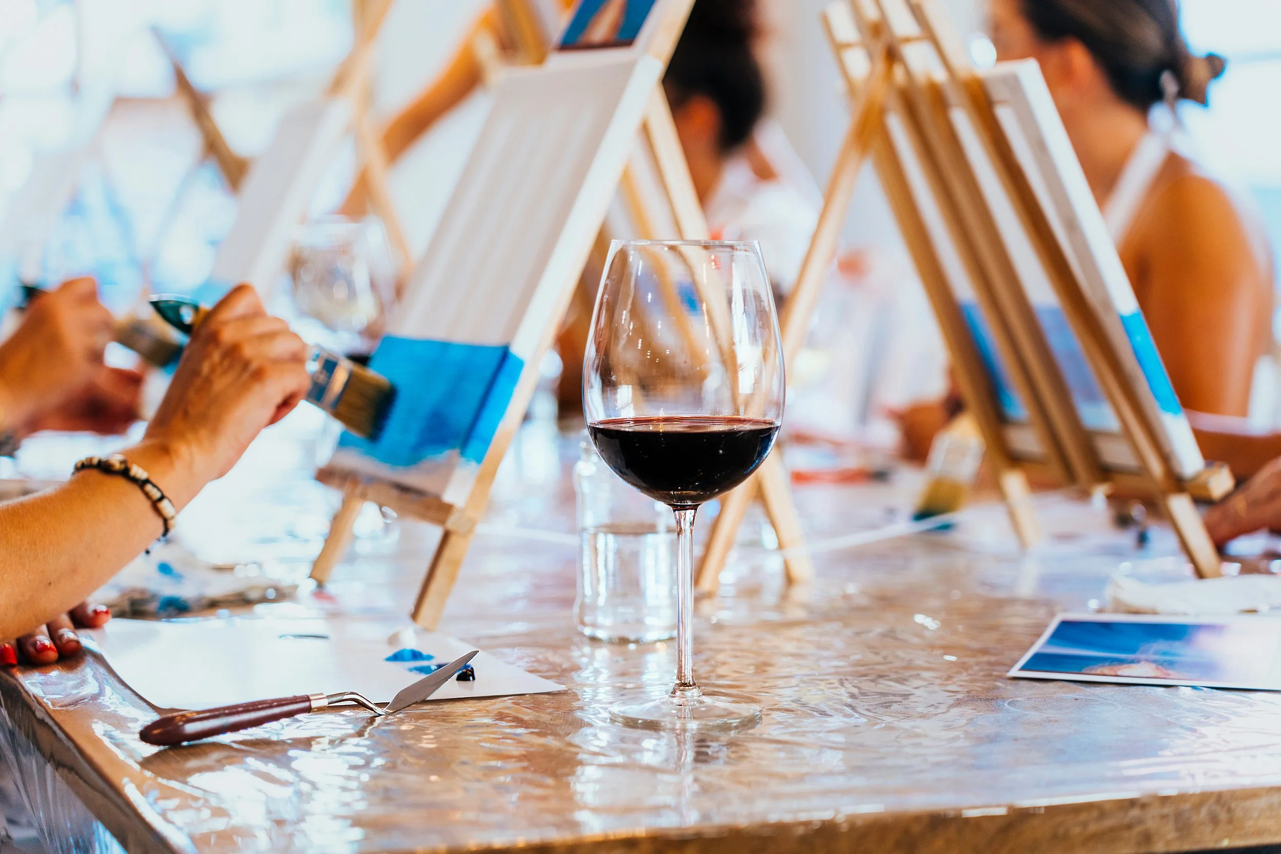 A glass of red wine on a table in front of people painting on canvases set on easels.