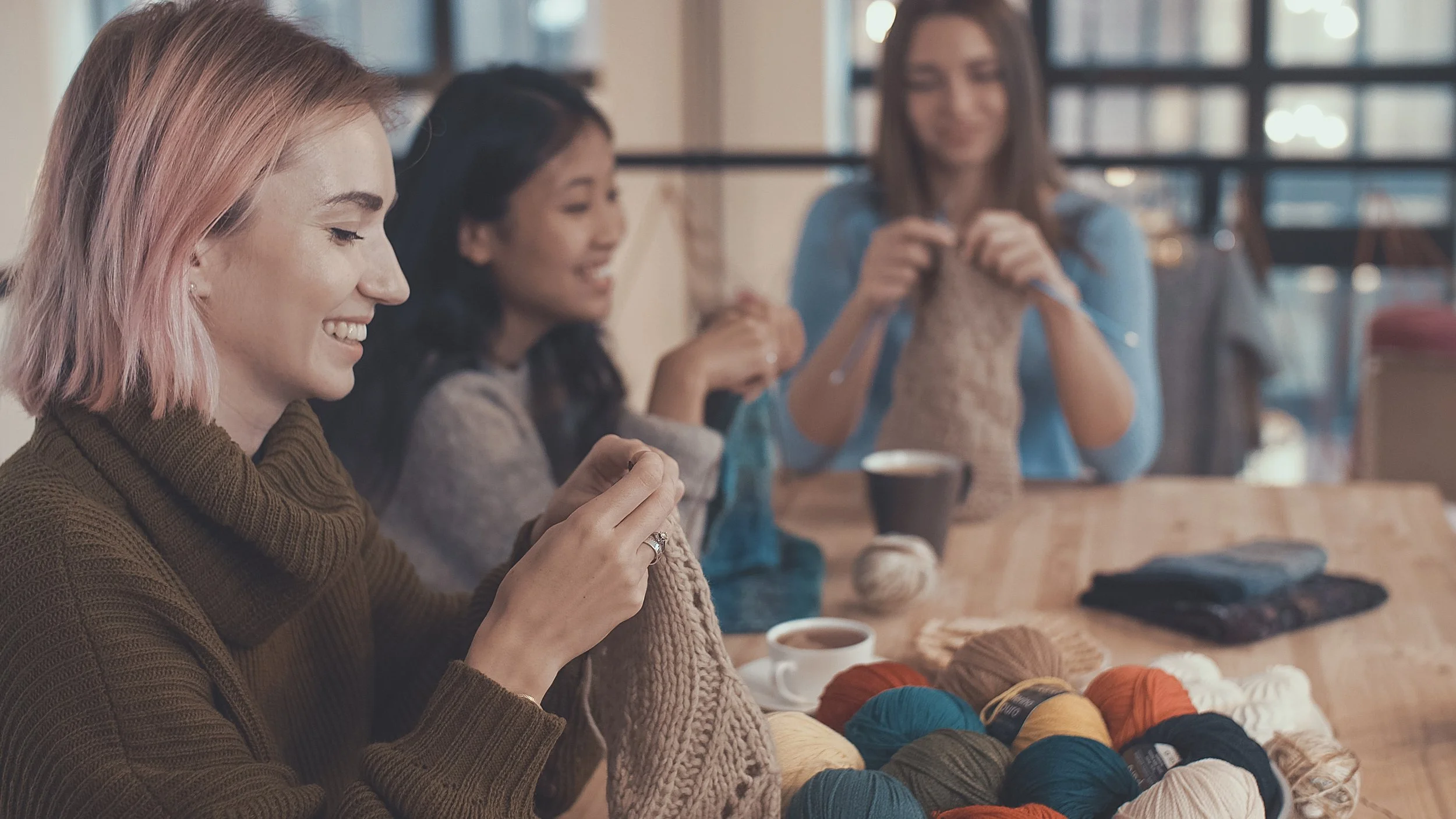Four women sitting at a wooden table knitting and chatting with balls of yarn and finished knitting projects in front of them.