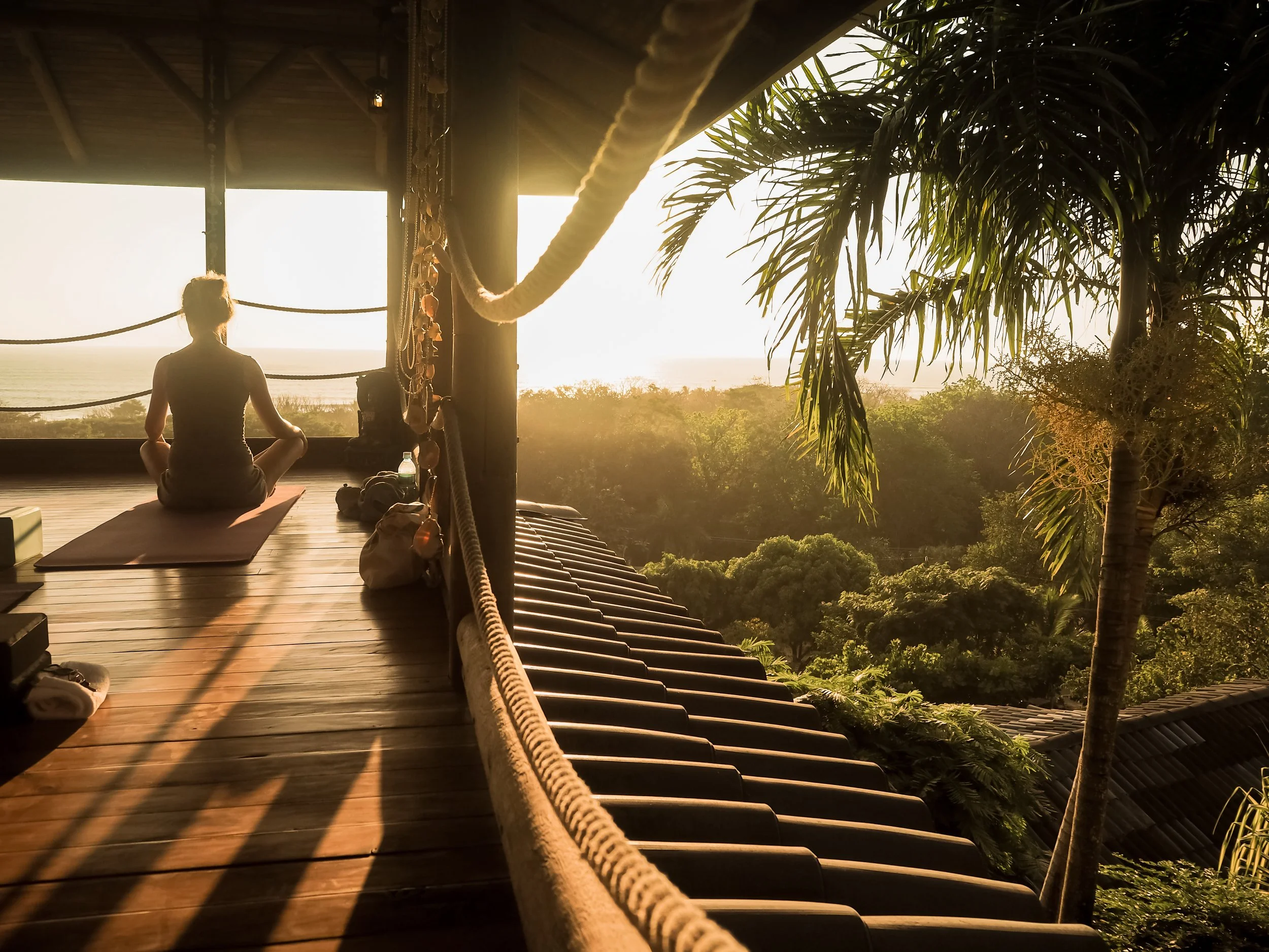 A woman practicing yoga on a wooden deck at sunrise, overlooking a lush tropical landscape with trees and palm trees.