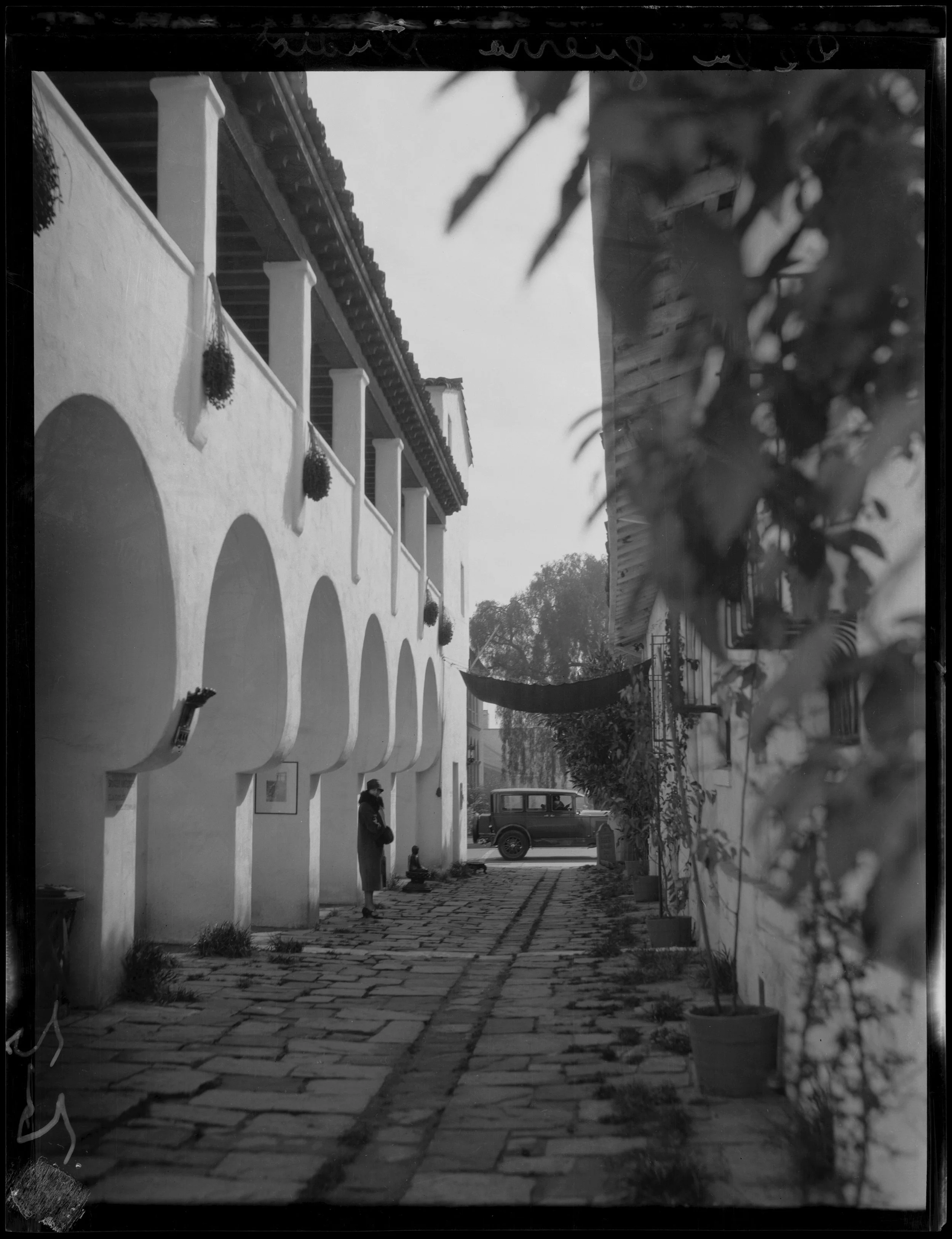 A black and white photo of a cobblestone street in front of a white building with arches and hanging potted plants. A vintage car is parked in the background, with a person standing near the wall. Plants are visible on the right side.