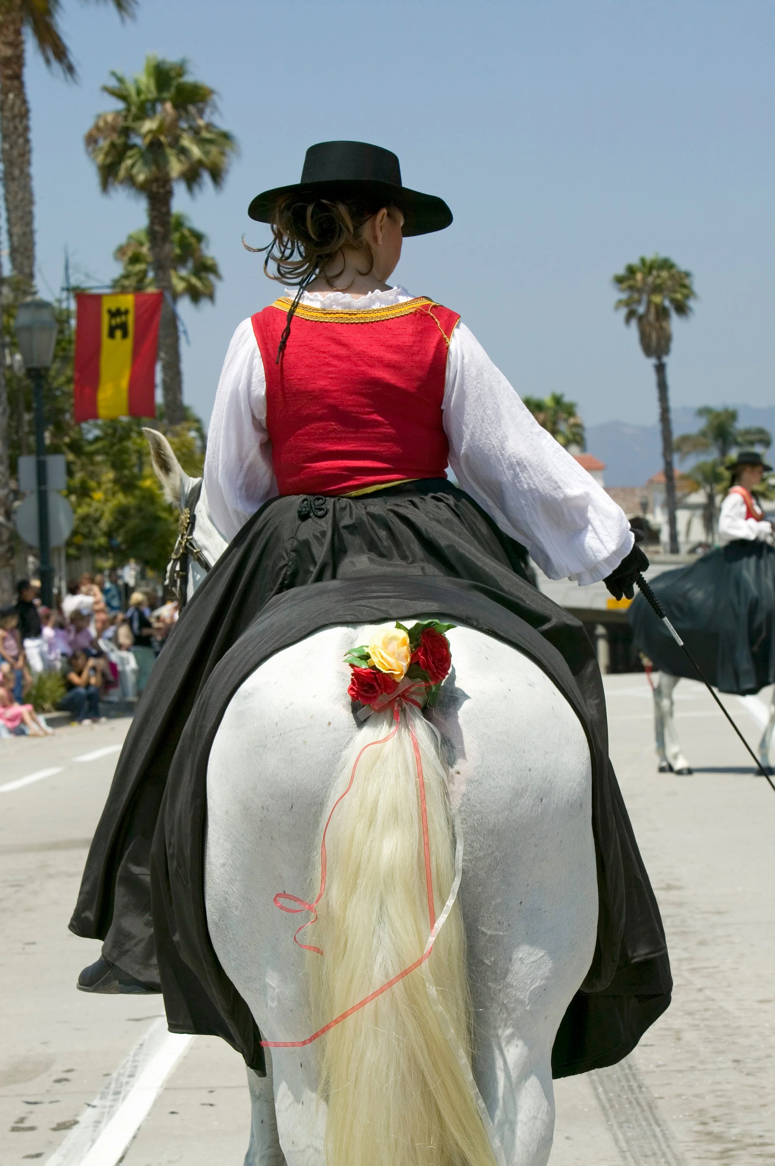 Woman in traditional dress riding a white horse in a parade with palm trees and spectators in the background.
