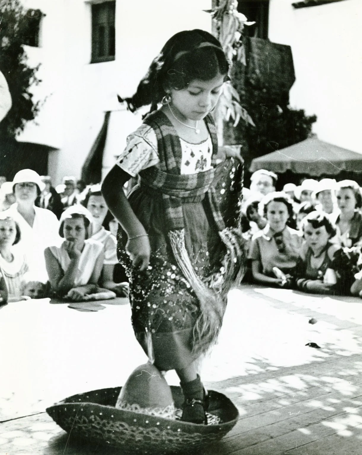 A young girl performing a traditional dance while standing on a circular prop, with an audience watching outdoors under umbrellas.