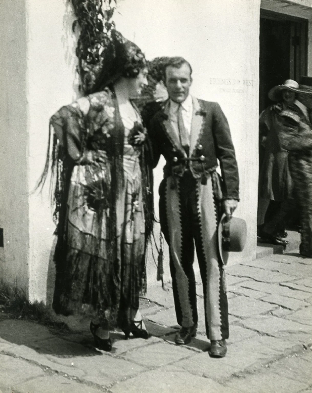 Black and white photo of a woman and a man dressed in traditional Mexican clothing, standing on a cobblestone street outside a building.