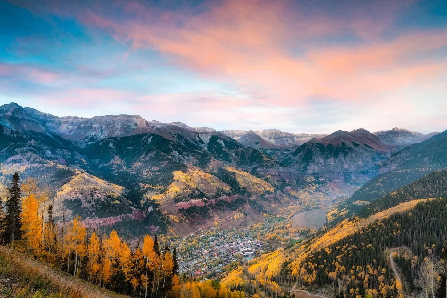Scenic view of a mountain valley during sunset with colorful autumn trees, a small town, and a winding river.