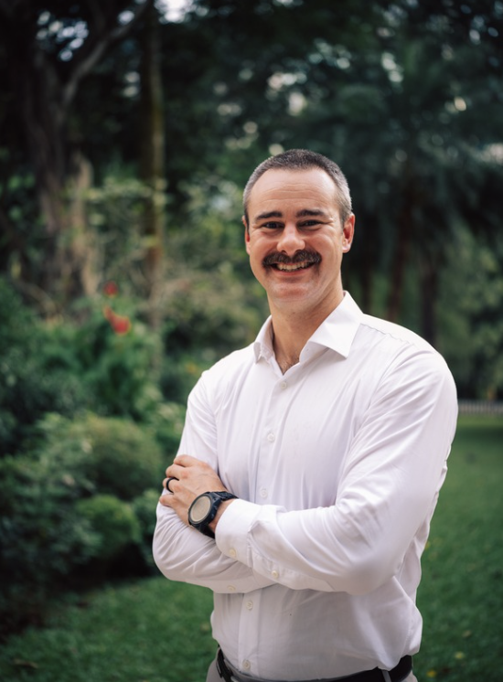 Man with a mustache smiling outdoors in a white shirt with arms crossed, standing in a garden or park with green trees and bushes in the background.