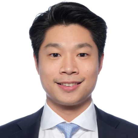 Headshot of a young man with black hair, smiling, dressed in a dark suit and light blue tie, against a white background.