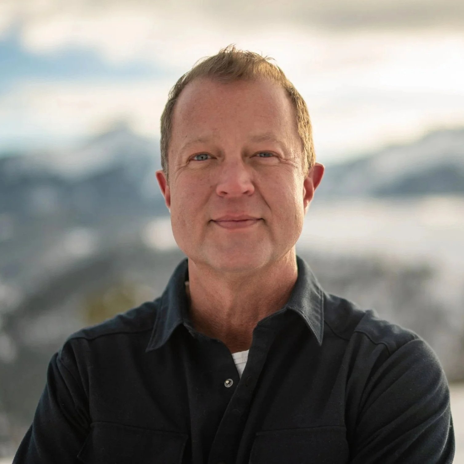 A smiling man with crossed arms standing outdoors in a park with trees and greenery.