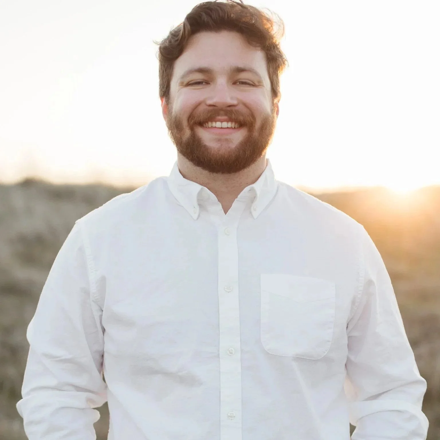 Smiling man with brown hair and beard wearing a white button-up shirt, outdoors during sunset.