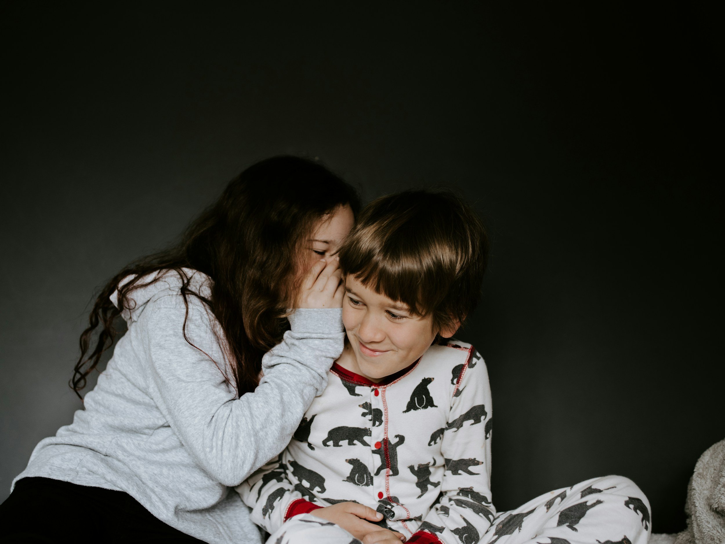 A girl with long curly hair whispering into a boy's ear, both smiling, against a dark background.
