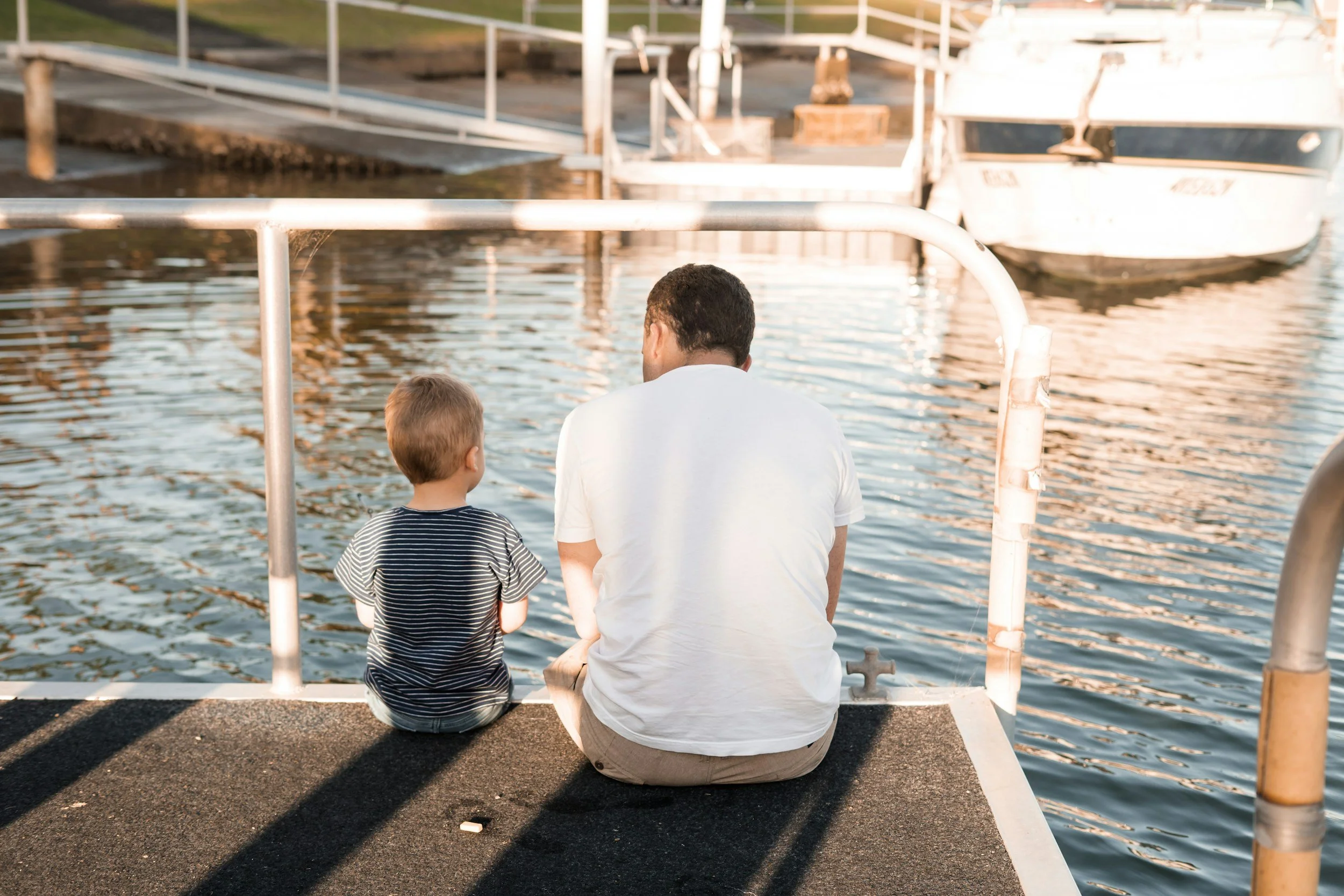 A man and a young boy sit side by side on a boat dock, facing the water, with boats moored nearby. The scene is bathed in warm sunlight.
