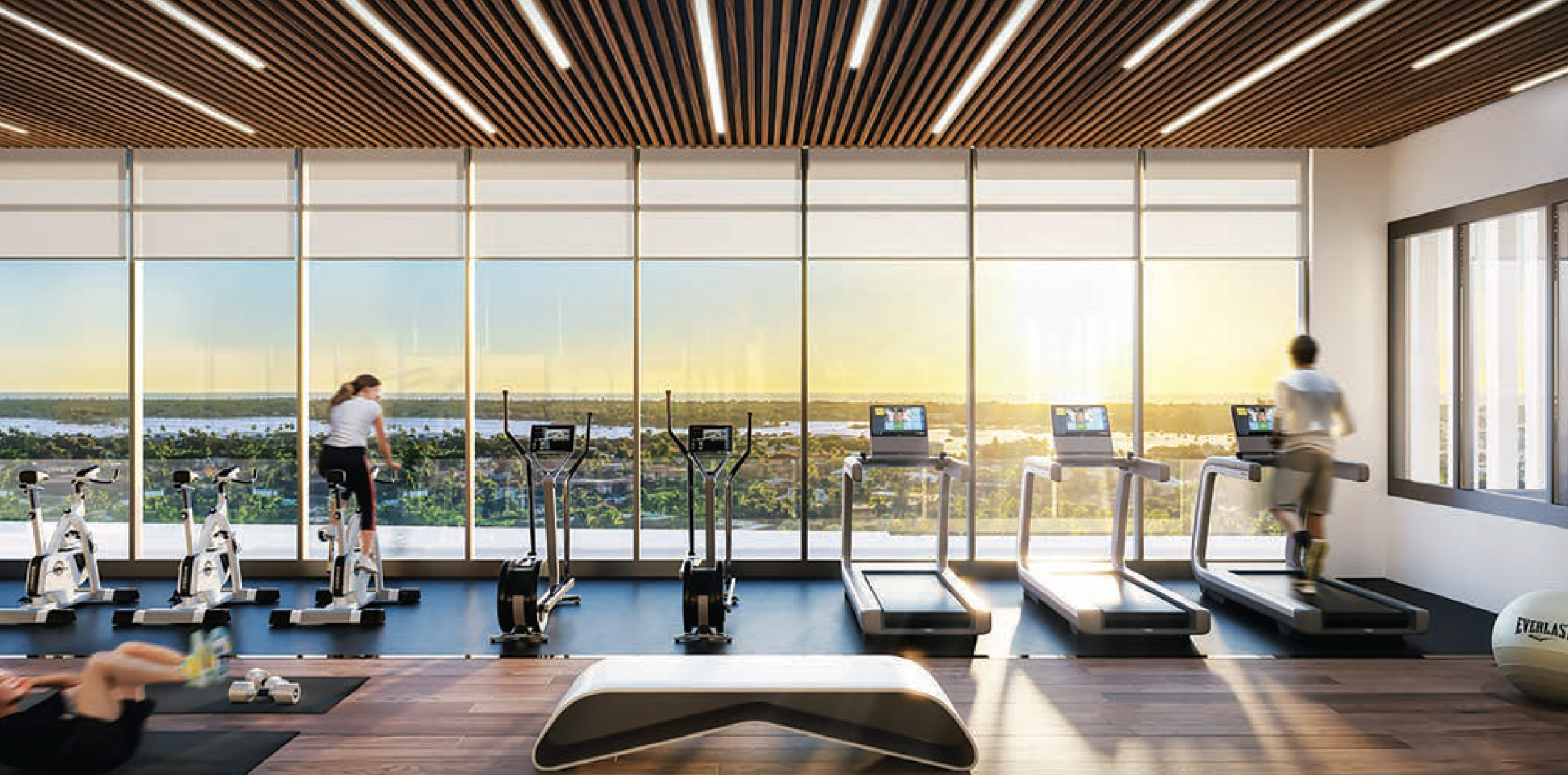 Gym with large windows showing a sunset over a cityscape, featuring exercise bikes, treadmills, and a person stretching on a mat.