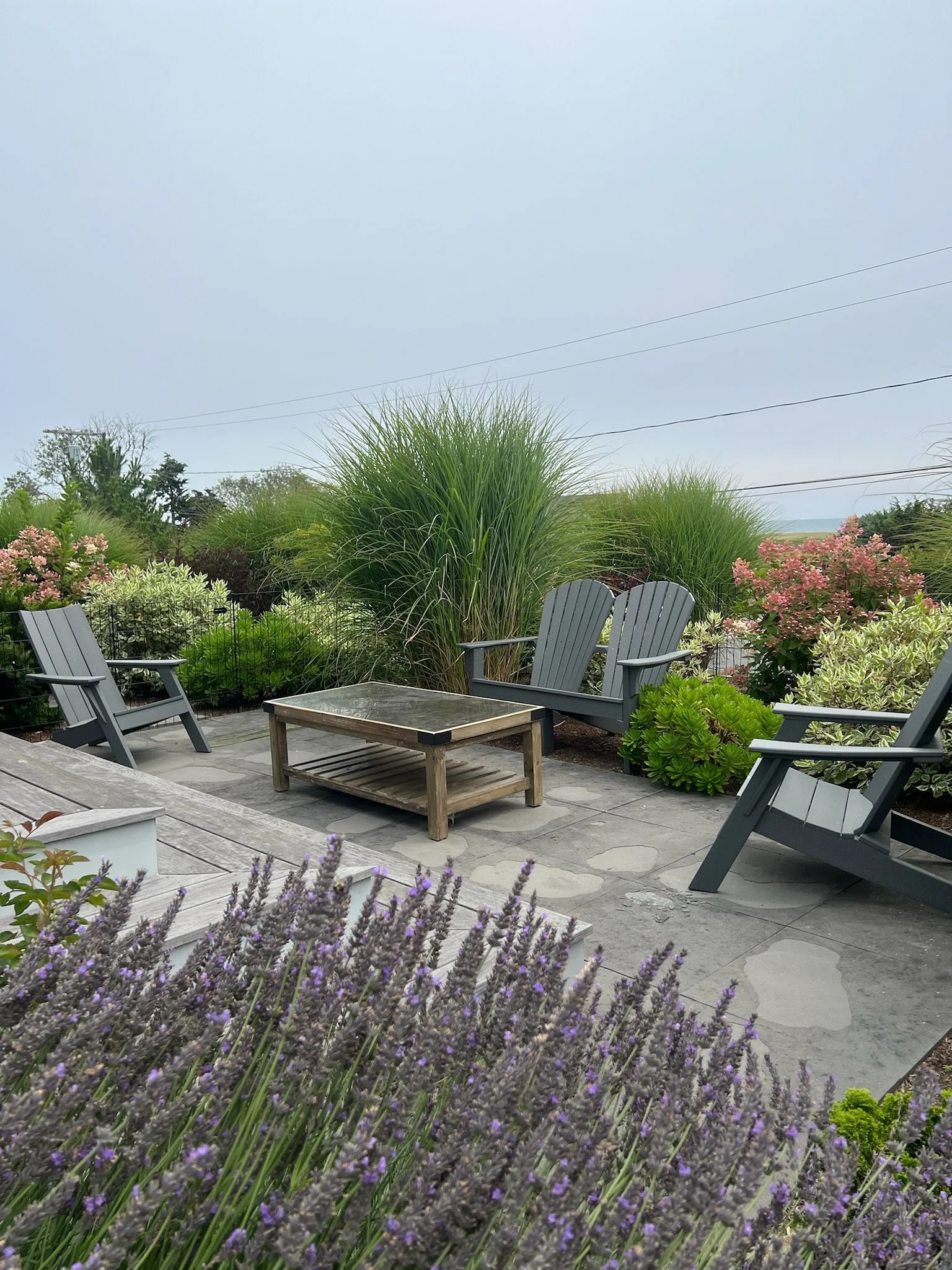 Outdoor patio with four gray Adirondack chairs around a square wooden table, surrounded by lush greenery and flowering plants, overcast sky in the background.