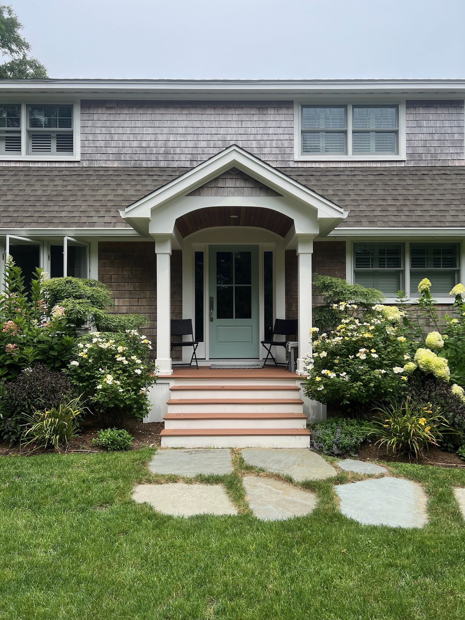 Front porch of a house with steps leading up, surrounded by green bushes and white flowers. The house has a covered entryway with white columns, a teal front door, and two black chairs on the porch.