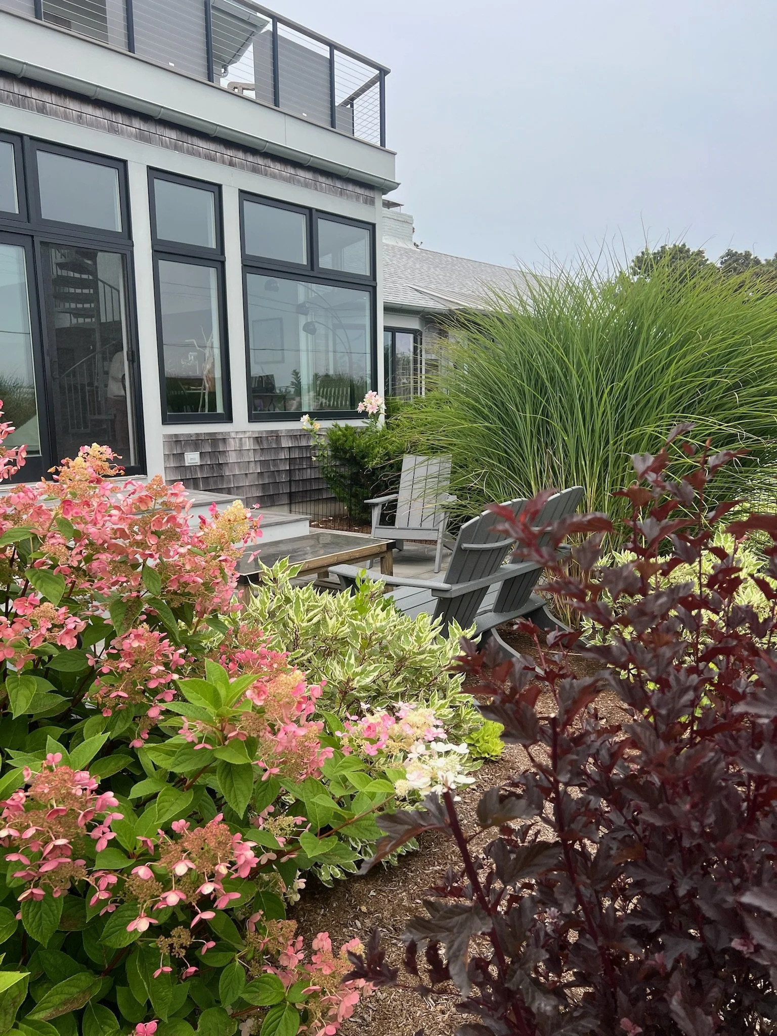 A backyard garden with pink and white flowering shrubs, a large ornamental grass, two gray Adirondack chairs, and a small wooden table in front of a house with large windows and a second-floor balcony.