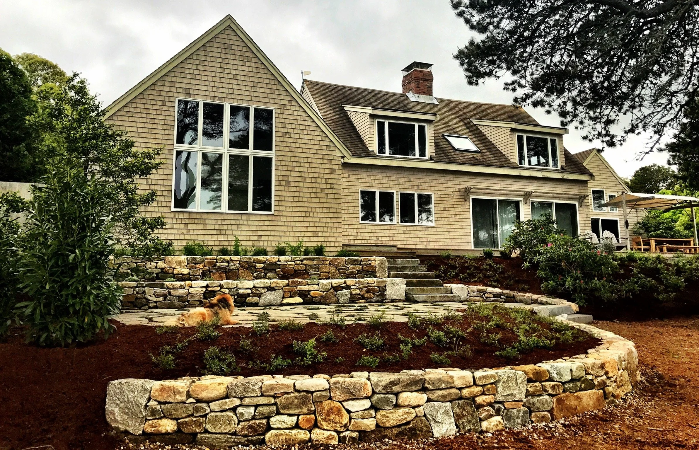 A large house with multiple windows and a brick chimney, surrounded by landscaped gardening with a stone retaining wall and a dog lying on the ground, and trees overhead.