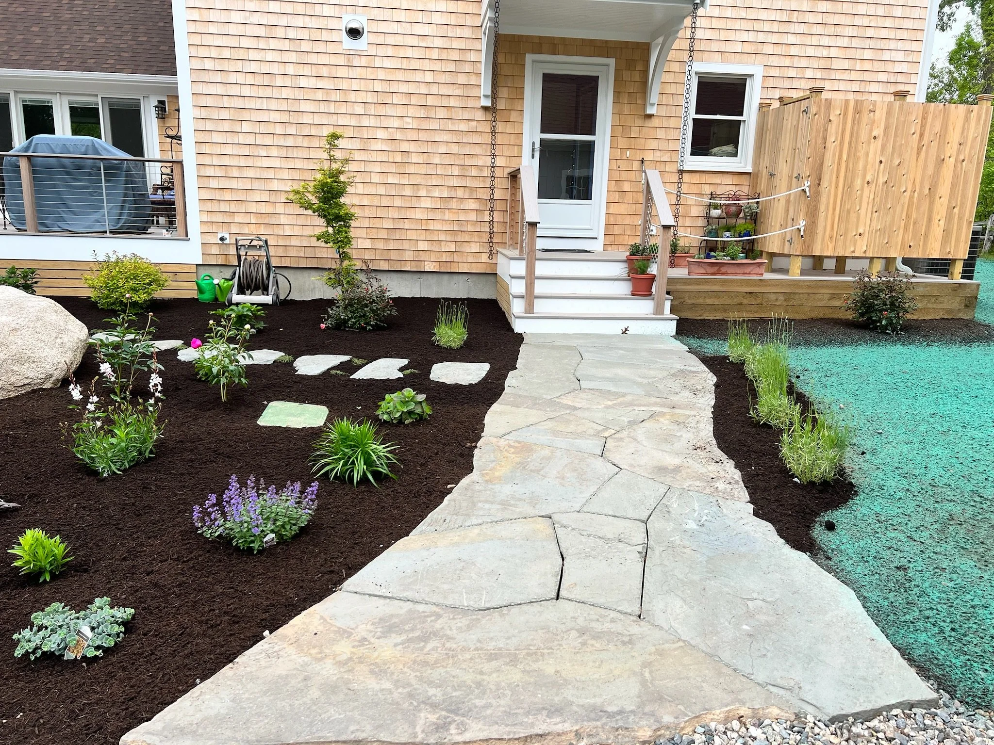 A landscaped backyard with a stone pathway leading to a house with a small porch and potted plants. The yard features freshly tilled soil, small bushes, and flowering plants, with a wooden deck and a privacy fence on one side.